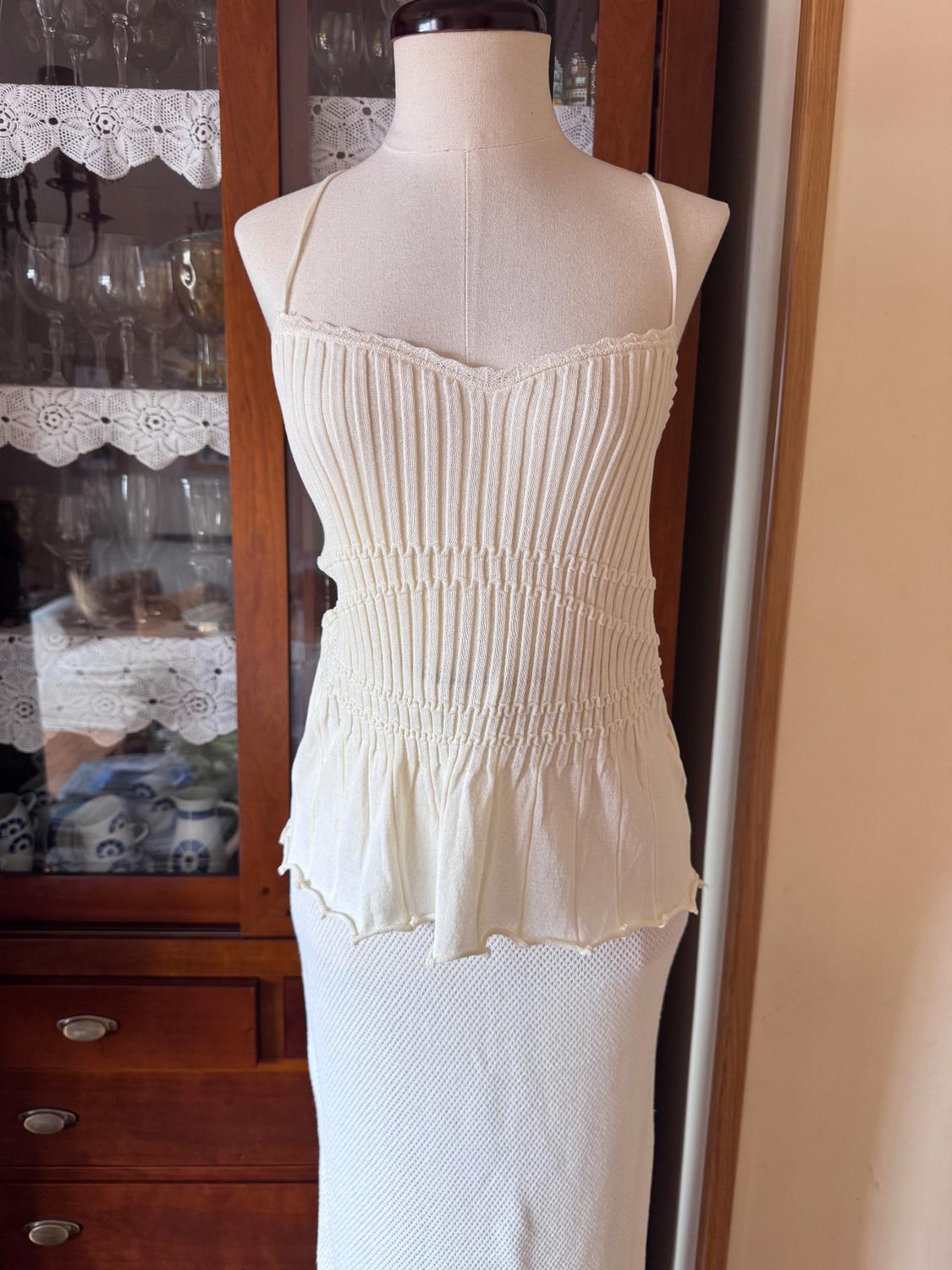 Mannequin in a white slip dress with a pleated bodice detail, in front of a wooden cabinet with glassware.