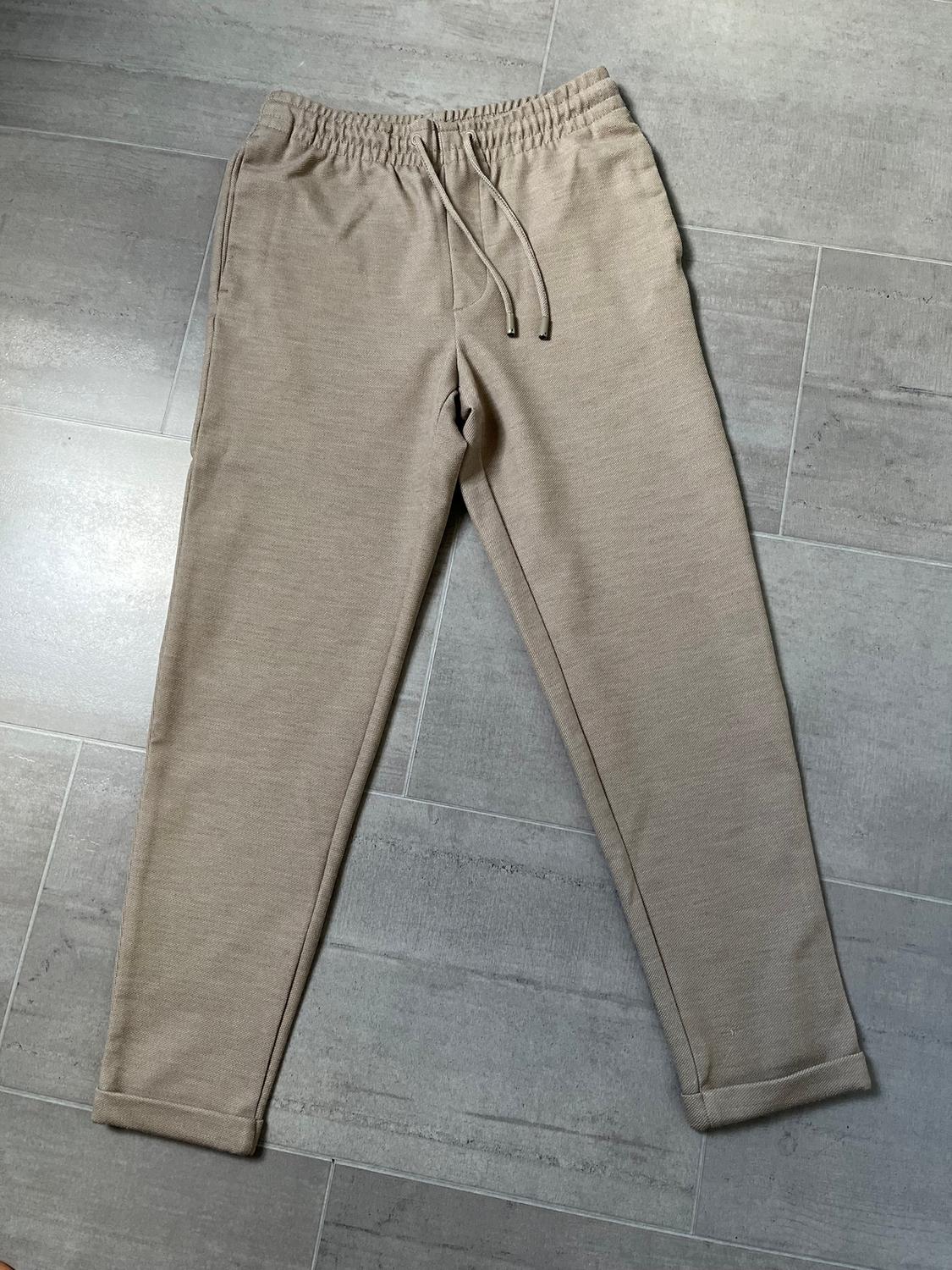 Beige tracksuit bottoms with an elasticated drawstring waist, placed on a grey tiled floor.