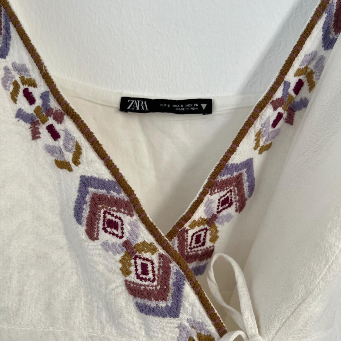Detail of a white blouse with geometric embroidery in shades of purple, pink and mustard. Visible label: "ZARA EUR S USA S MEX 26 MADE IN INDIA".