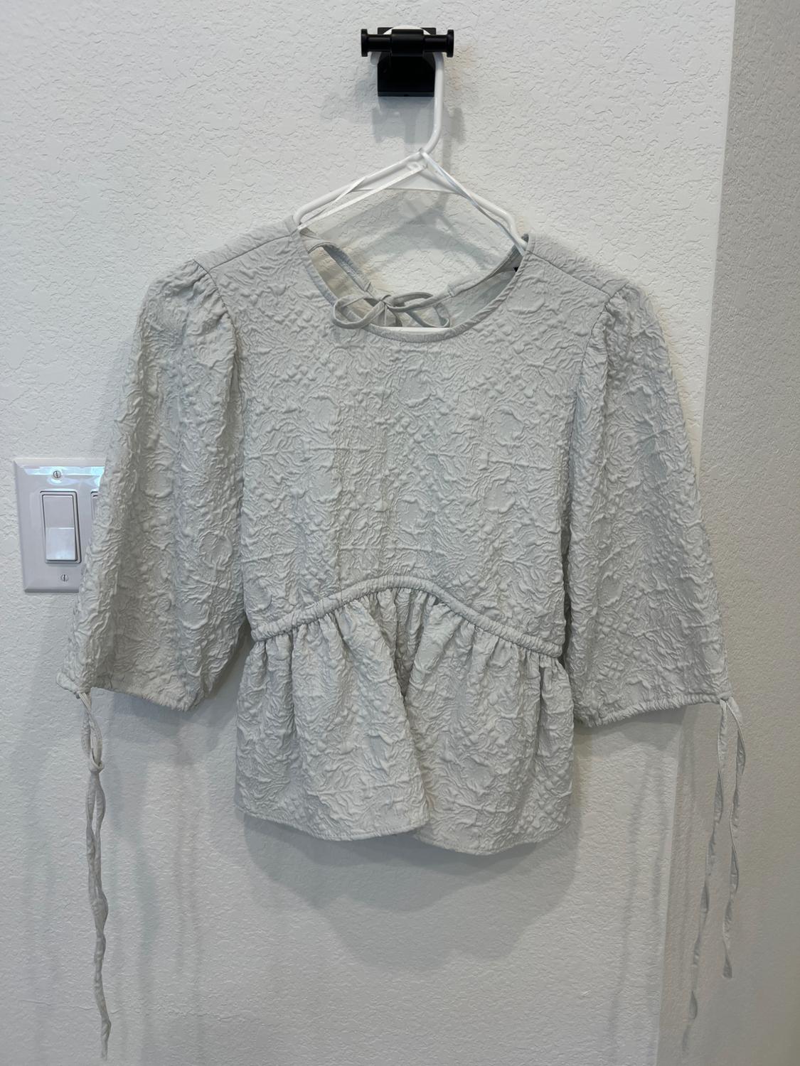 Light gray textured blouse with puff sleeves and cuff ties, hanging on a white hanger against a white wall.