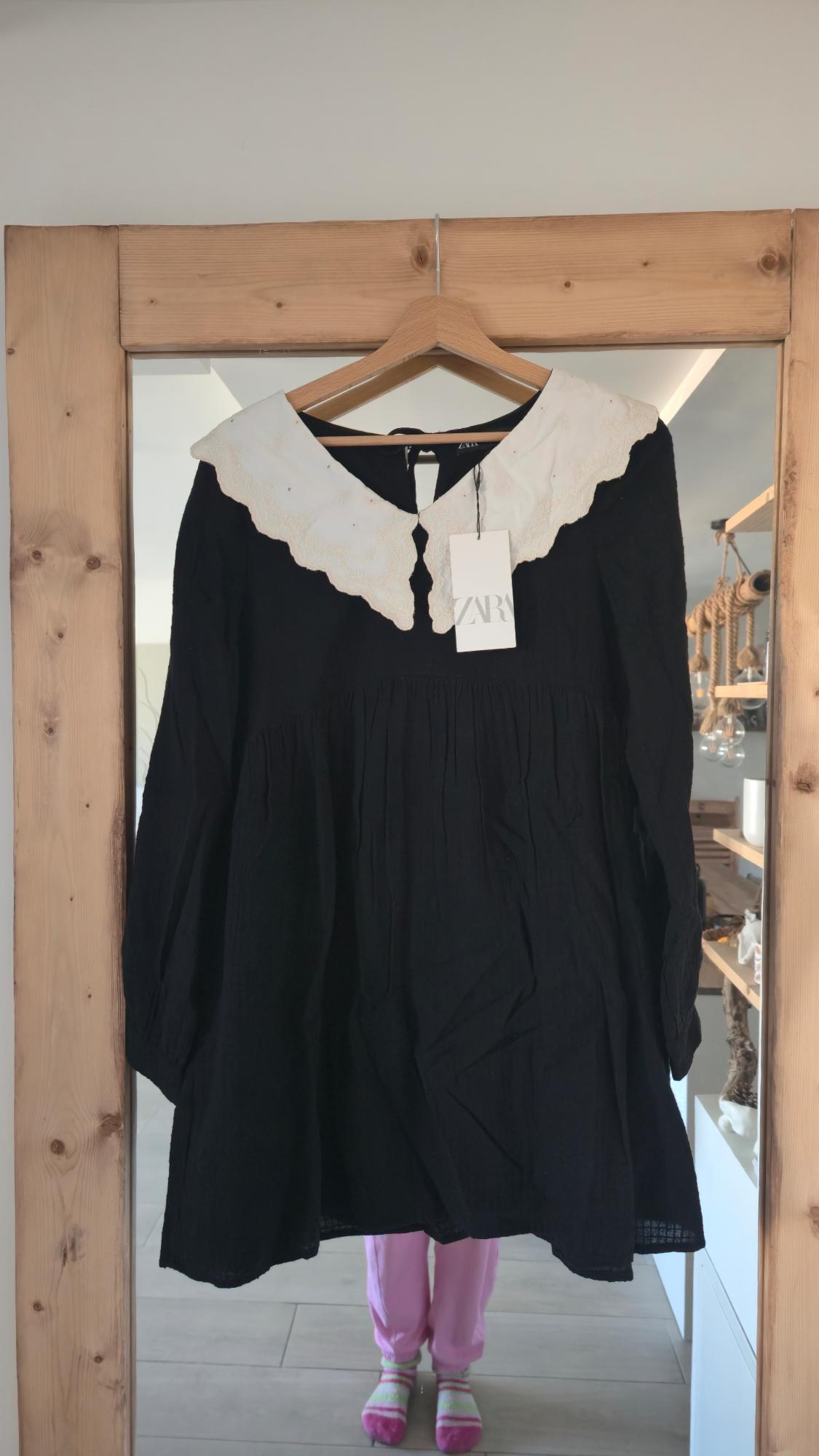 Black dress with white collar hanging on a wooden mirror. Reflection of a person in pink trousers and striped socks. "ZARA" label.
