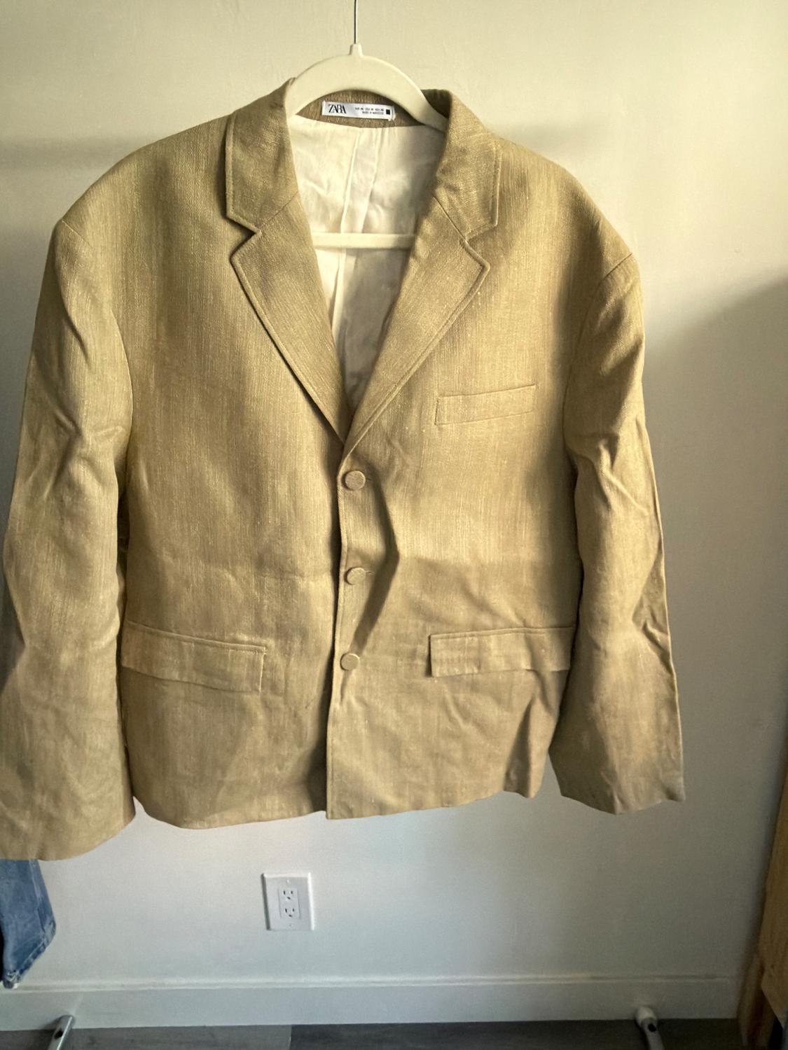 Beige jacket hanging on a white hanger, with lapels and three front buttons, against a light background. Visible label on the collar.