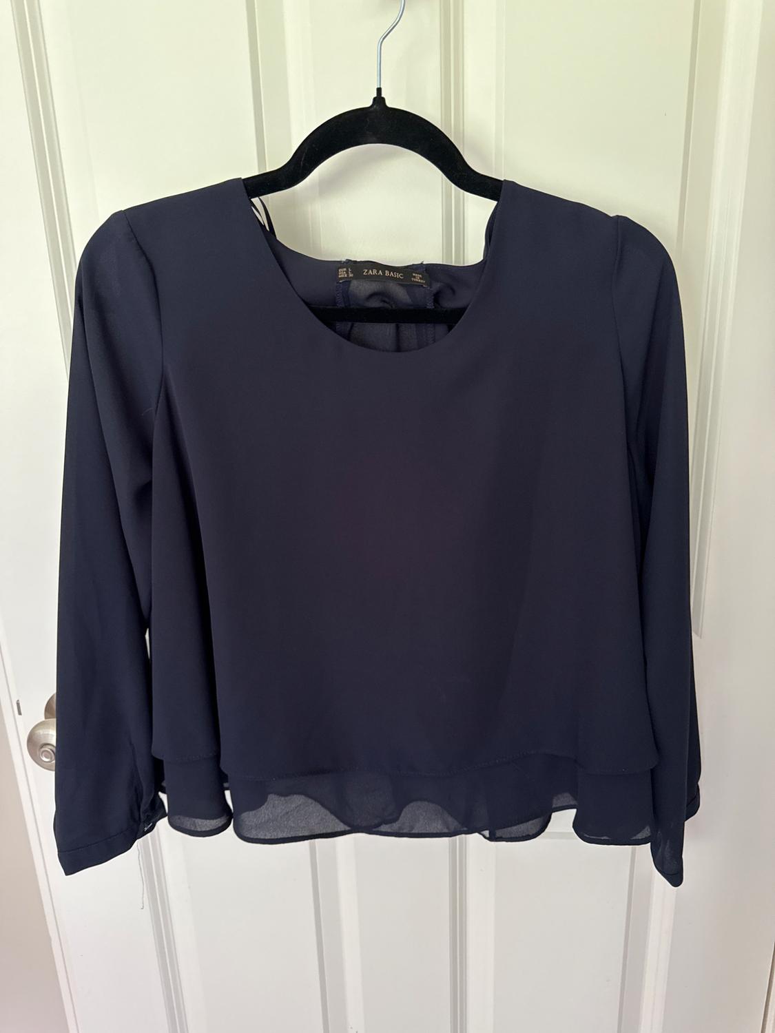 Navy blue long-sleeve blouse with a layered hem hanging on a white door. The label reads ZARA BASIC.