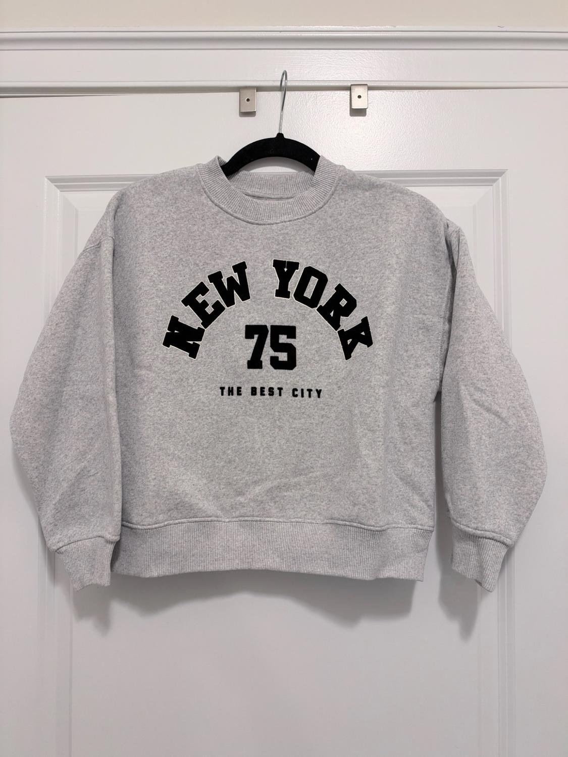 Light gray sweatshirt hanging on a white door. In the center, the black text
NEW YORK 75 THE BEST CITY stands out.