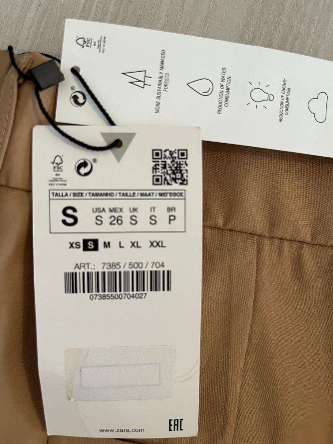 Beige clothing tag with size S information, barcode, and text: "More sustainably managed forests, reduction of water consumption, reduction of energy consumption".