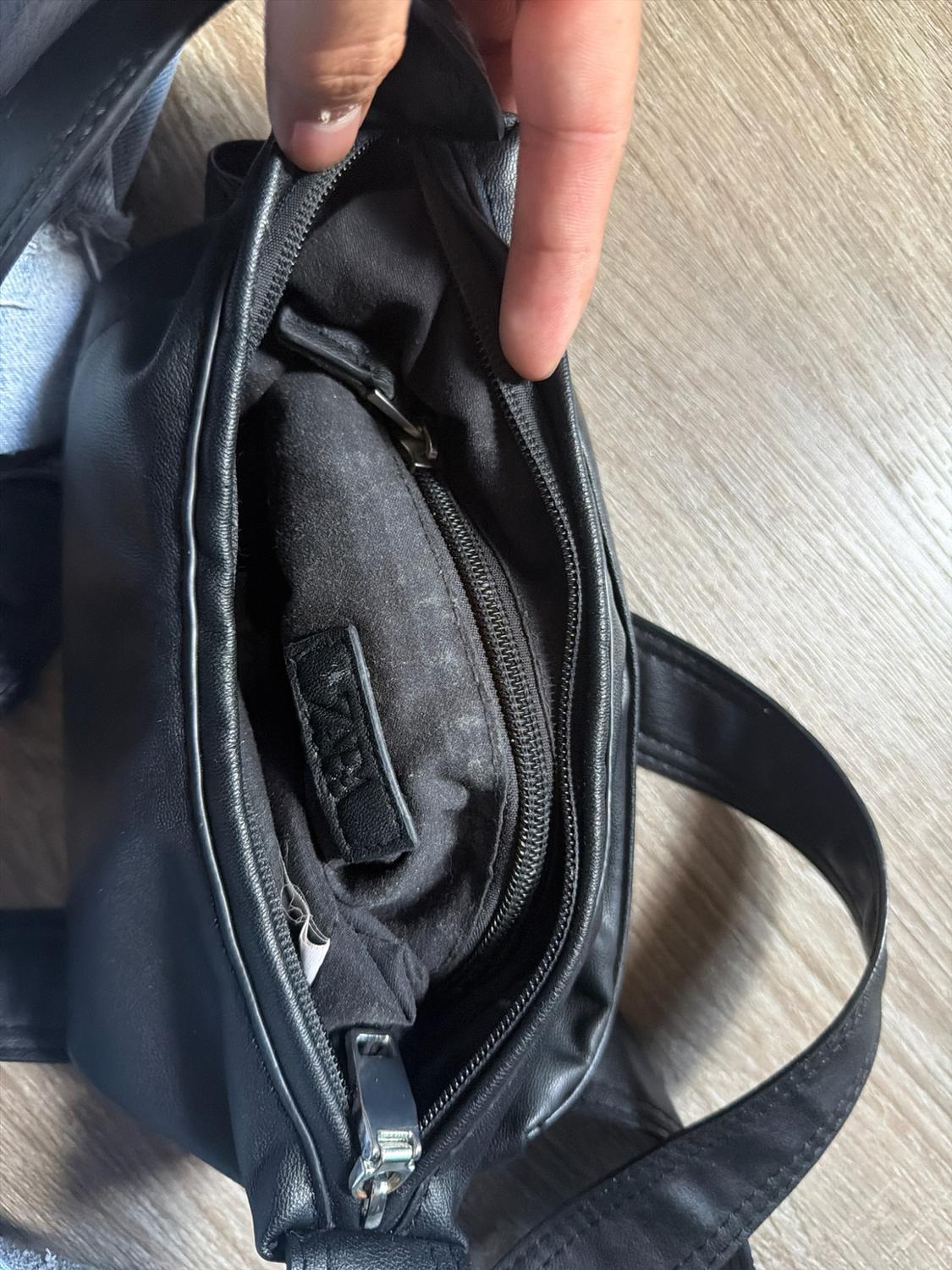 A hand opens a black handbag on a wooden floor. Inside, the lining, a zipper, and a tag with the text ZARA are visible.