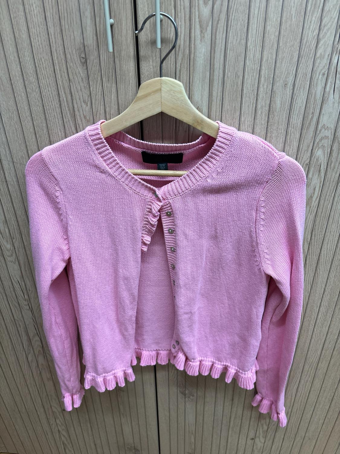 Pink knitted cardigan with buttons and ruffles, hanging on a hanger against a wood panel background.