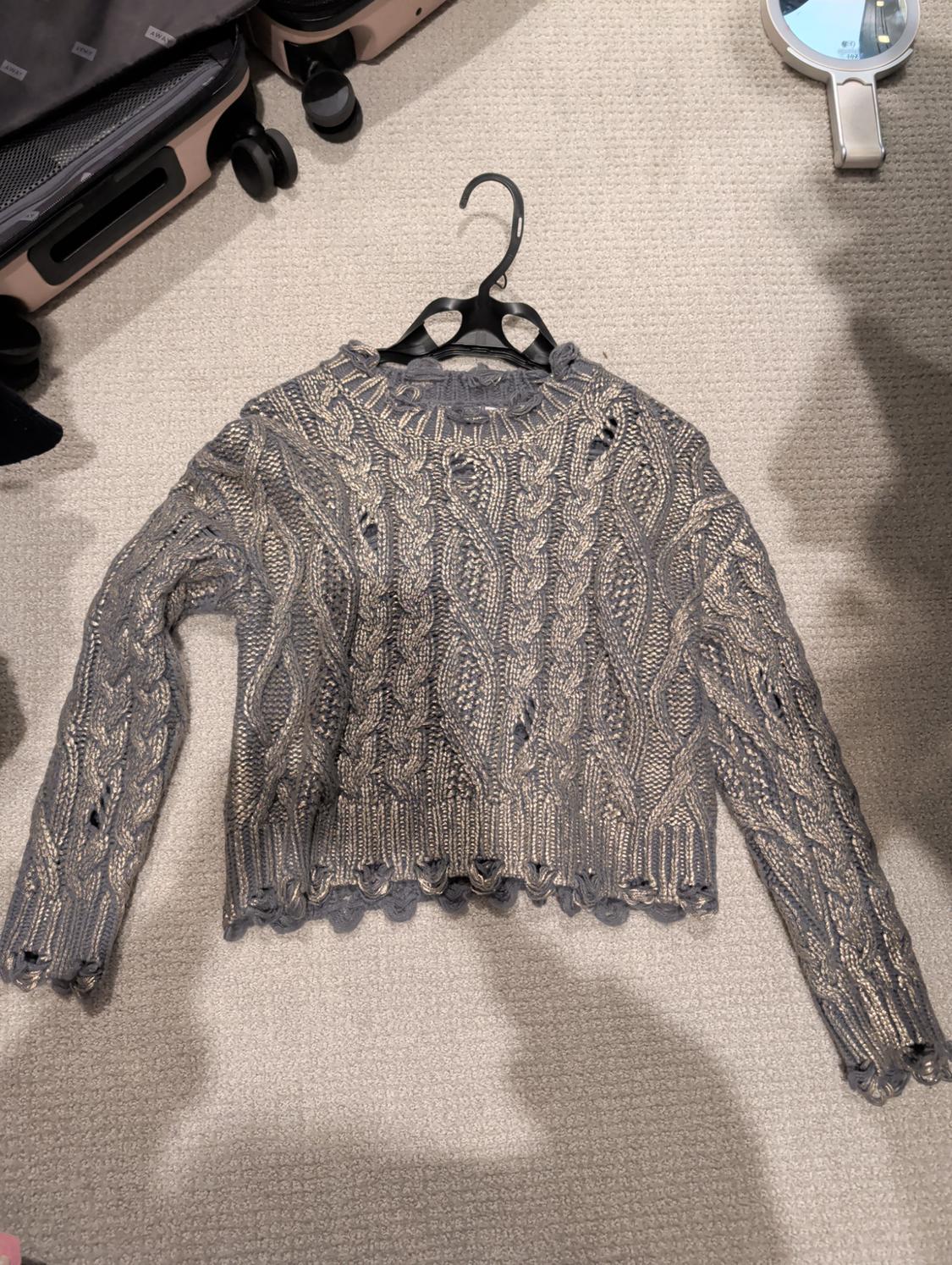 Grey knitted jumper with cable knit design and distressed details, hanging on a hanger over a beige carpet, next to suitcases and a mirror.