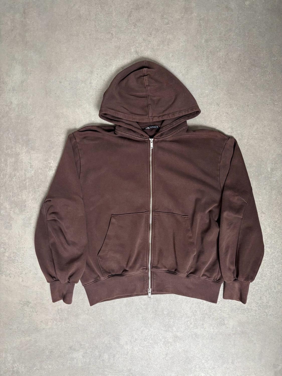 Brown hooded sweatshirt with a zip and front pockets, laid out on a textured grey surface. Casual style.