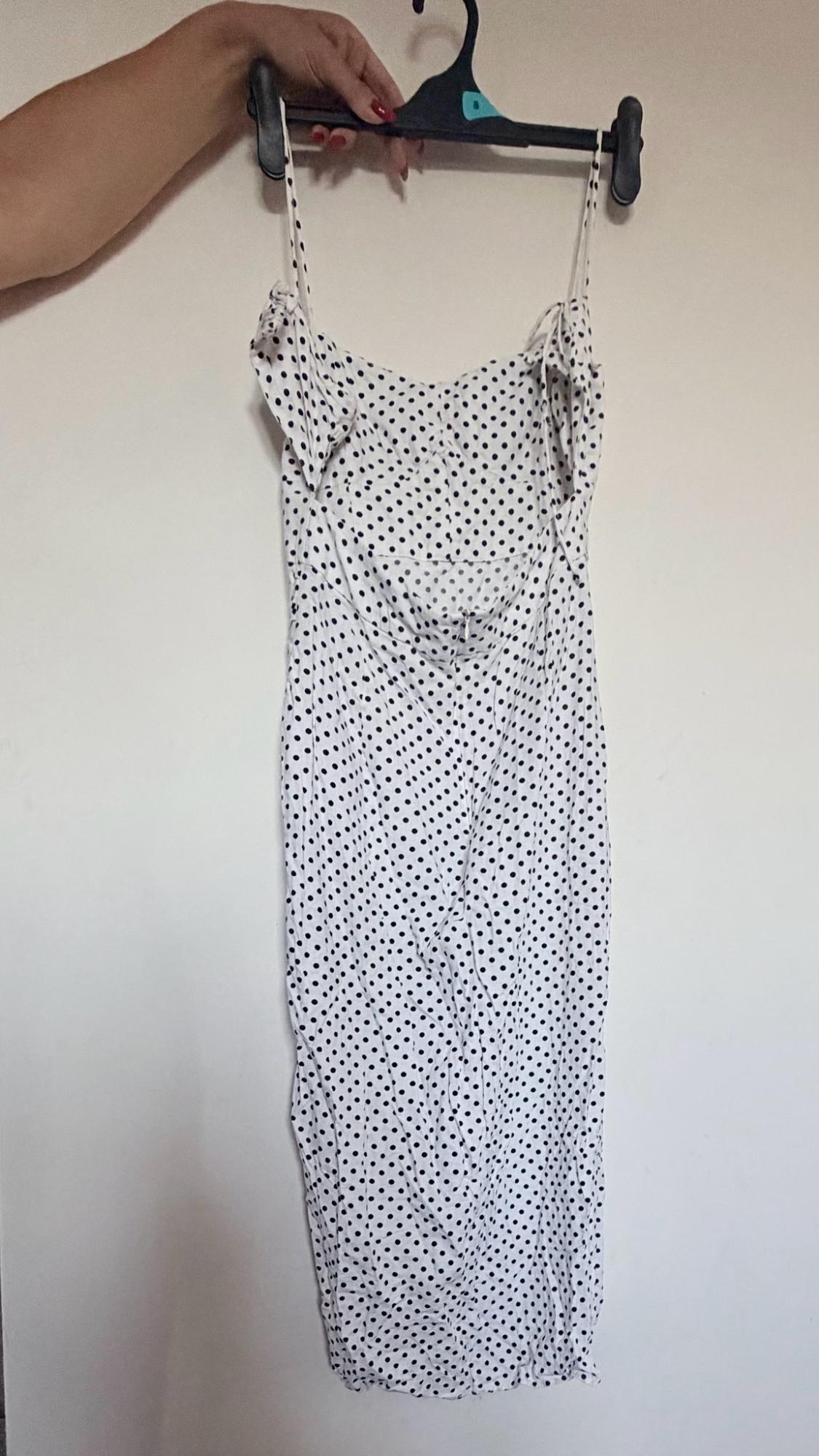 A hand holds a hanger with a white dress with black polka dots in front of a white wall. It has thin straps and an open back.