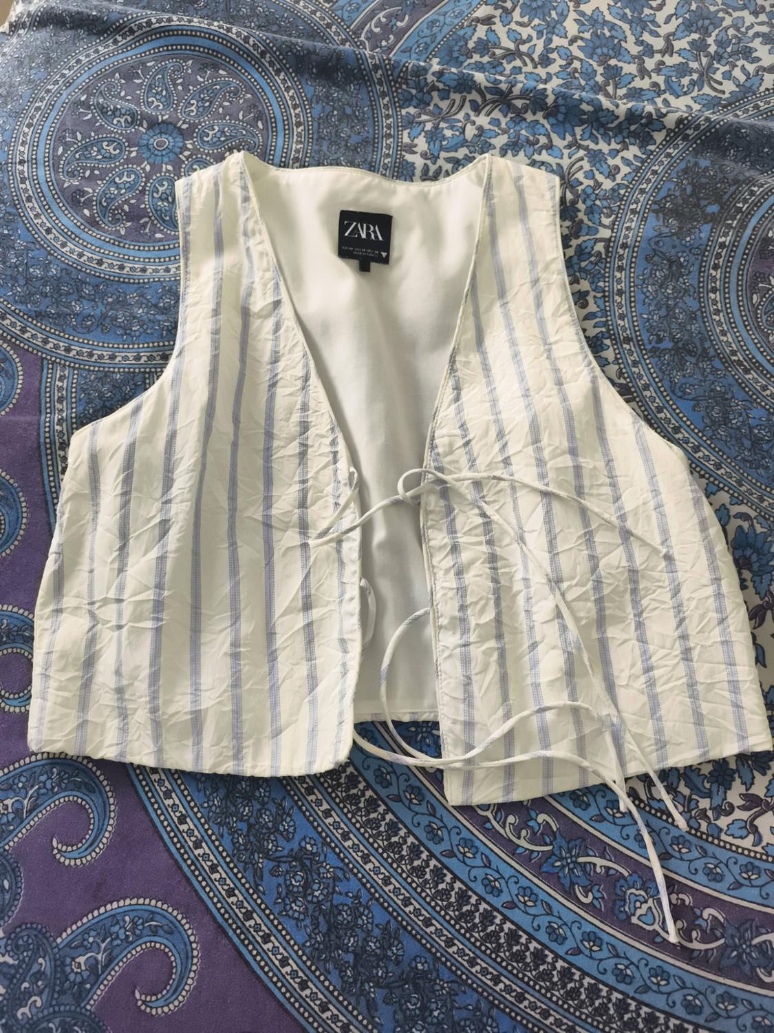 Creased white waistcoat with blue stripes and front ties, ZARA label, on a fabric with a blue and purple-toned print.