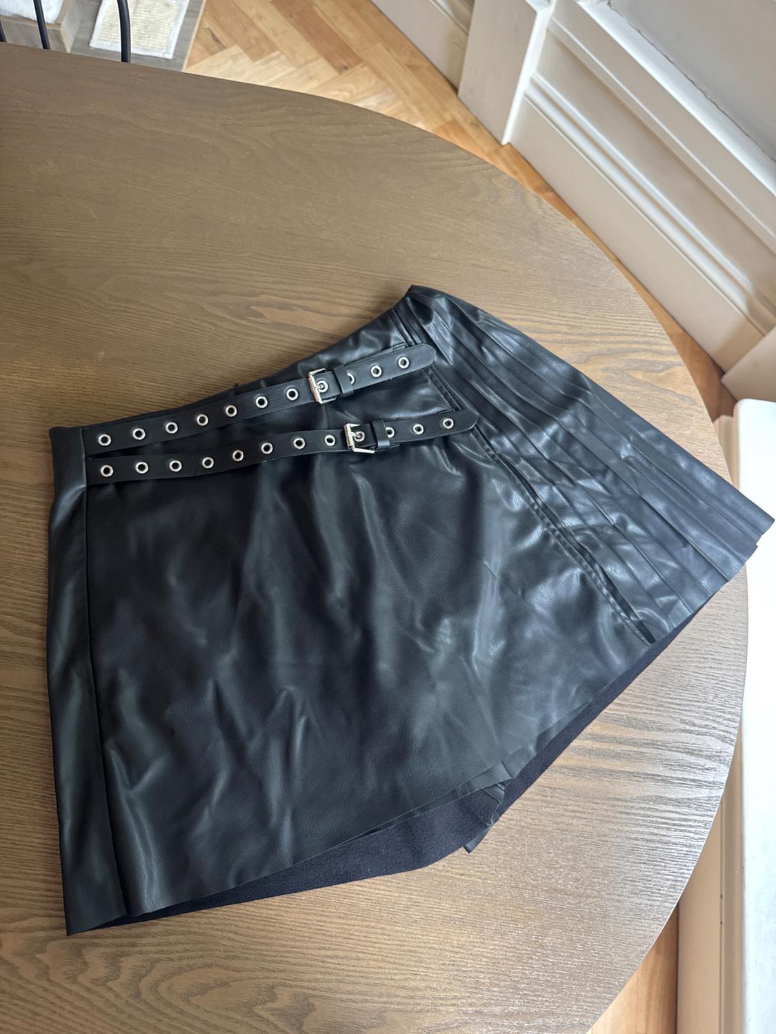 Black skirt made of synthetic material with buckles and metal eyelets, on a light wooden table in an indoor setting.