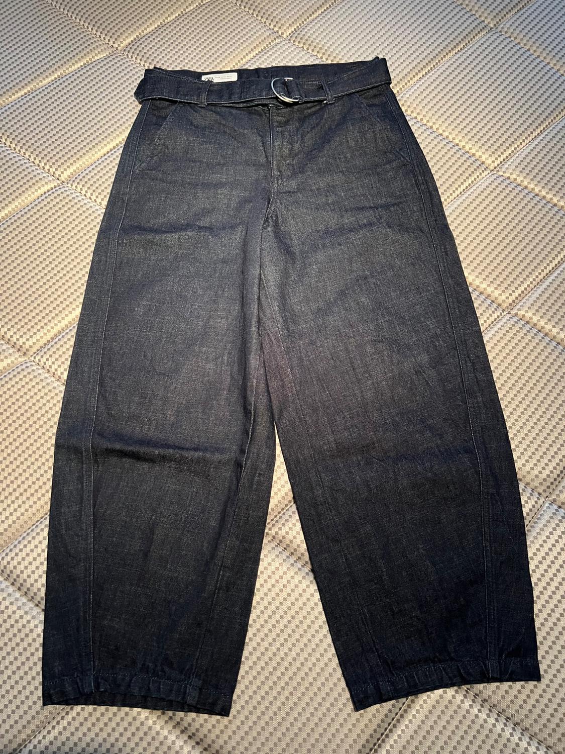 Dark denim trousers with a belt, placed on a padded surface with a geometric pattern.