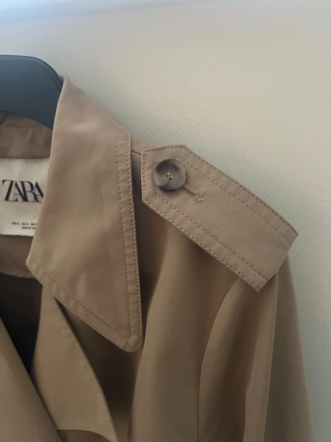 Beige trench coat hanging on a black hanger. Shoulder pad detail with button. Visible label with text "ZARA".