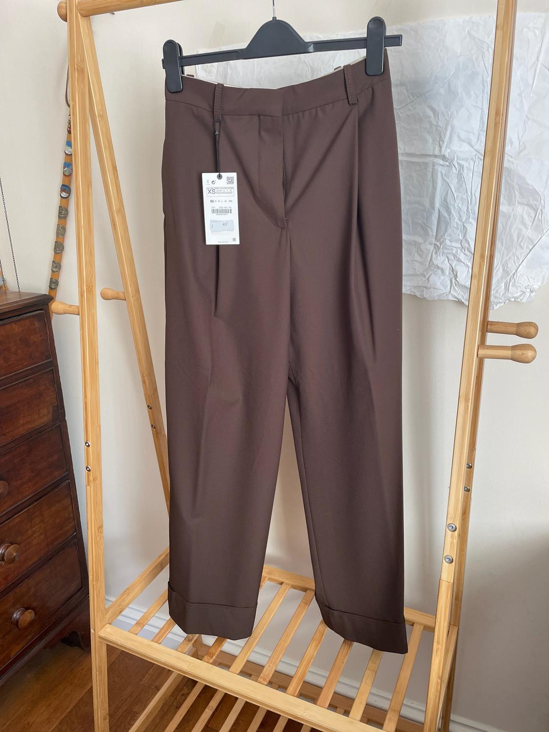 Brown pleated trousers hanging on a wooden clothes rail. Visible label with the text XS and 49. Background of a room with a wooden chest of drawers.
