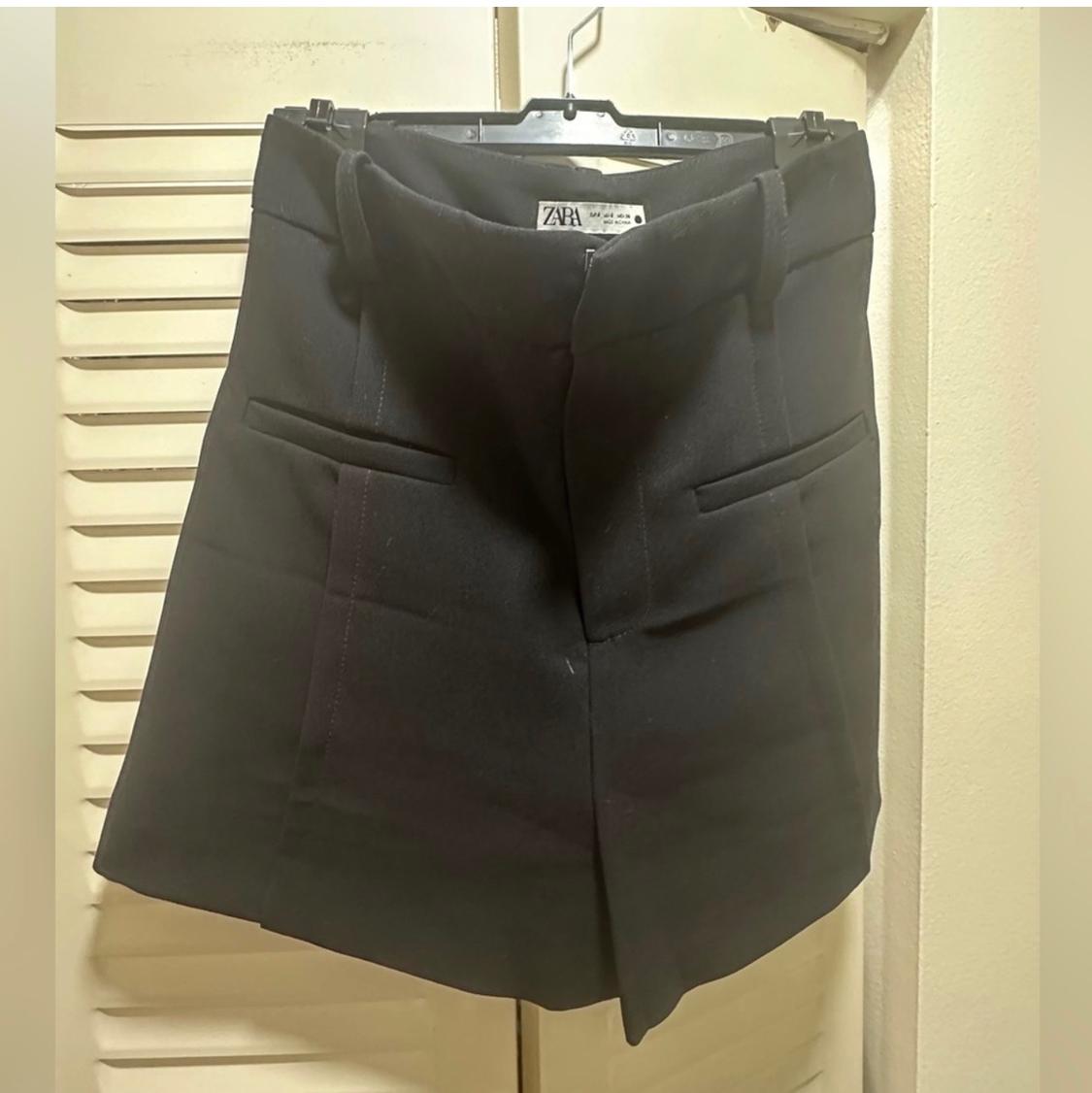 Black skirt hanging on a hanger, with front pockets and a visible Zara label. White door background.