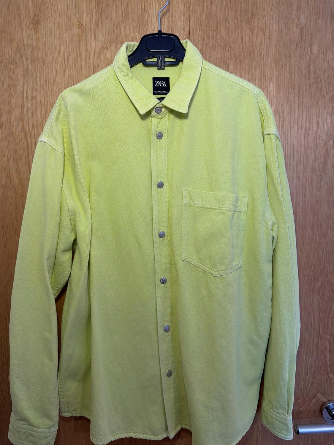 Light green long-sleeved shirt with metallic buttons and a chest pocket, hanging on a black hanger in front of a wooden door.