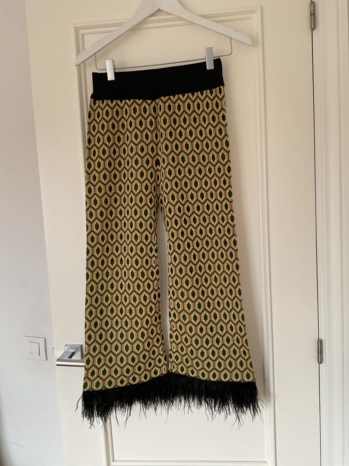 Trousers hanging on a hanger, with a yellow and black geometric print, and black fringes on the hem, over a white door.