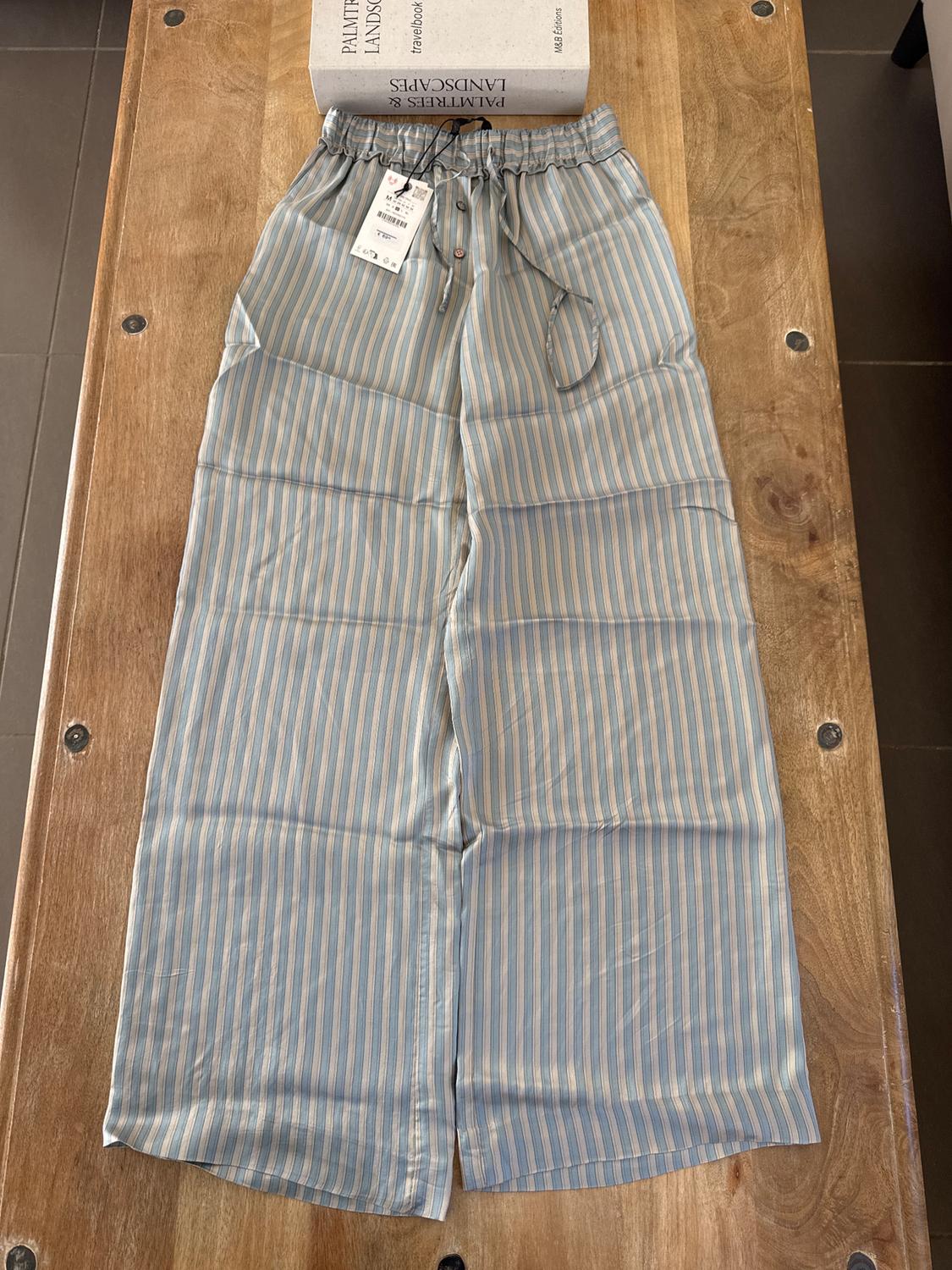 Blue and white striped trousers with an elasticated waist and tag, on a wooden table next to a travel book.