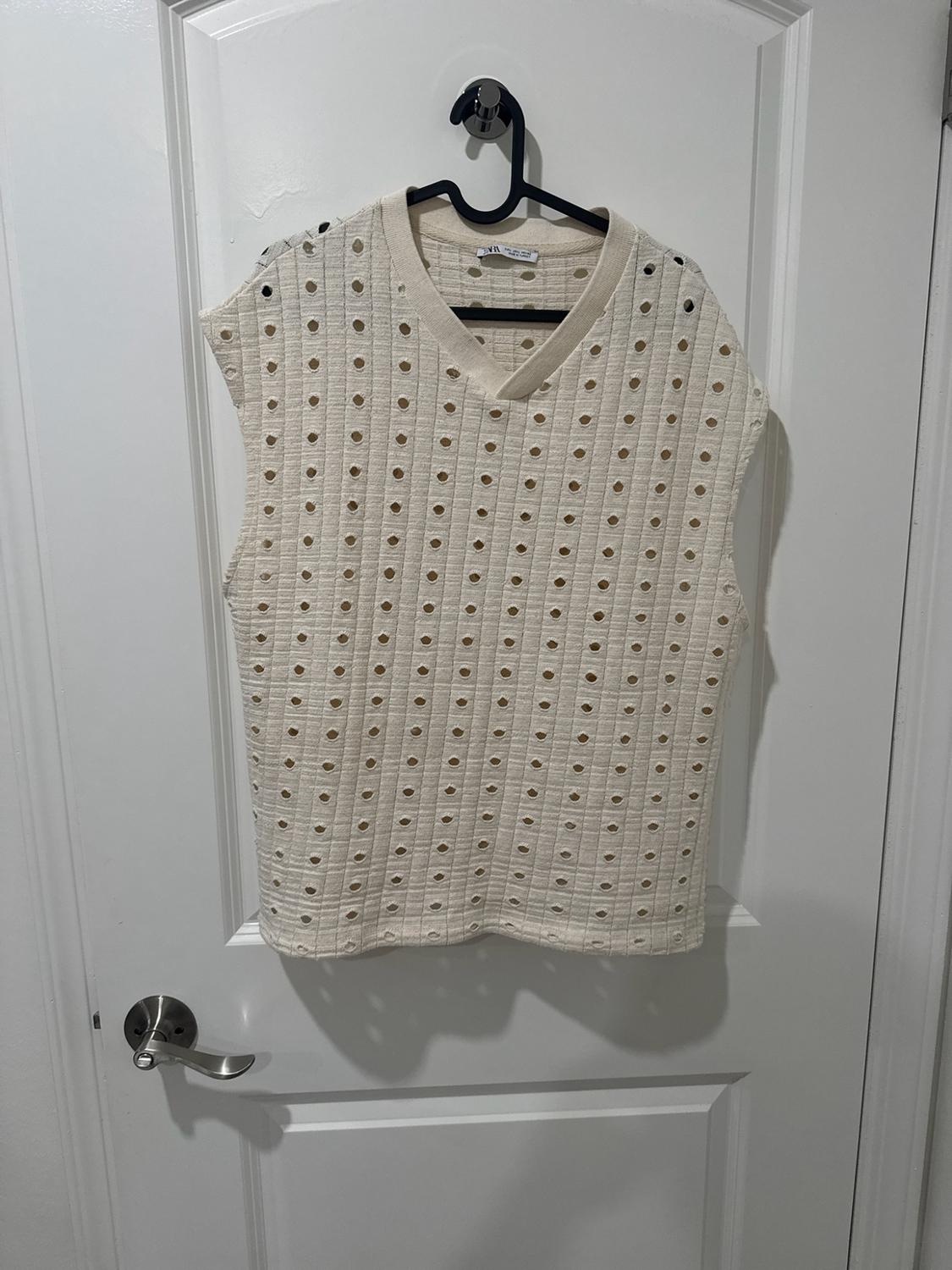 Cream-colored sleeveless vest with a V-neck and perforated texture, hanging on a black hanger over a white door.