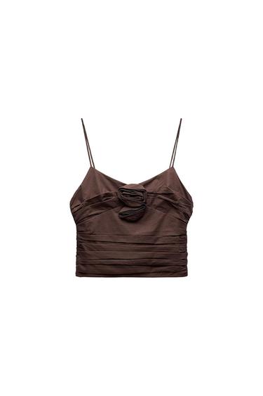 Brown crop top with spaghetti straps, ruched fabric, and a flower detail at the center of the neckline, on a white background.