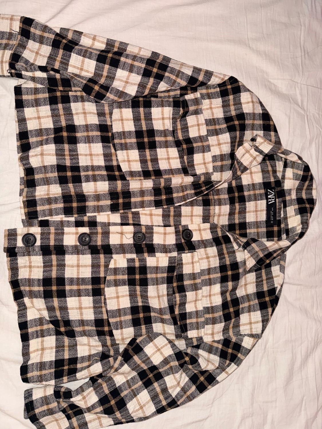 Plaid shirt in beige, black, and brown tones with black buttons, on a white surface. Visible label with text "ZARA".