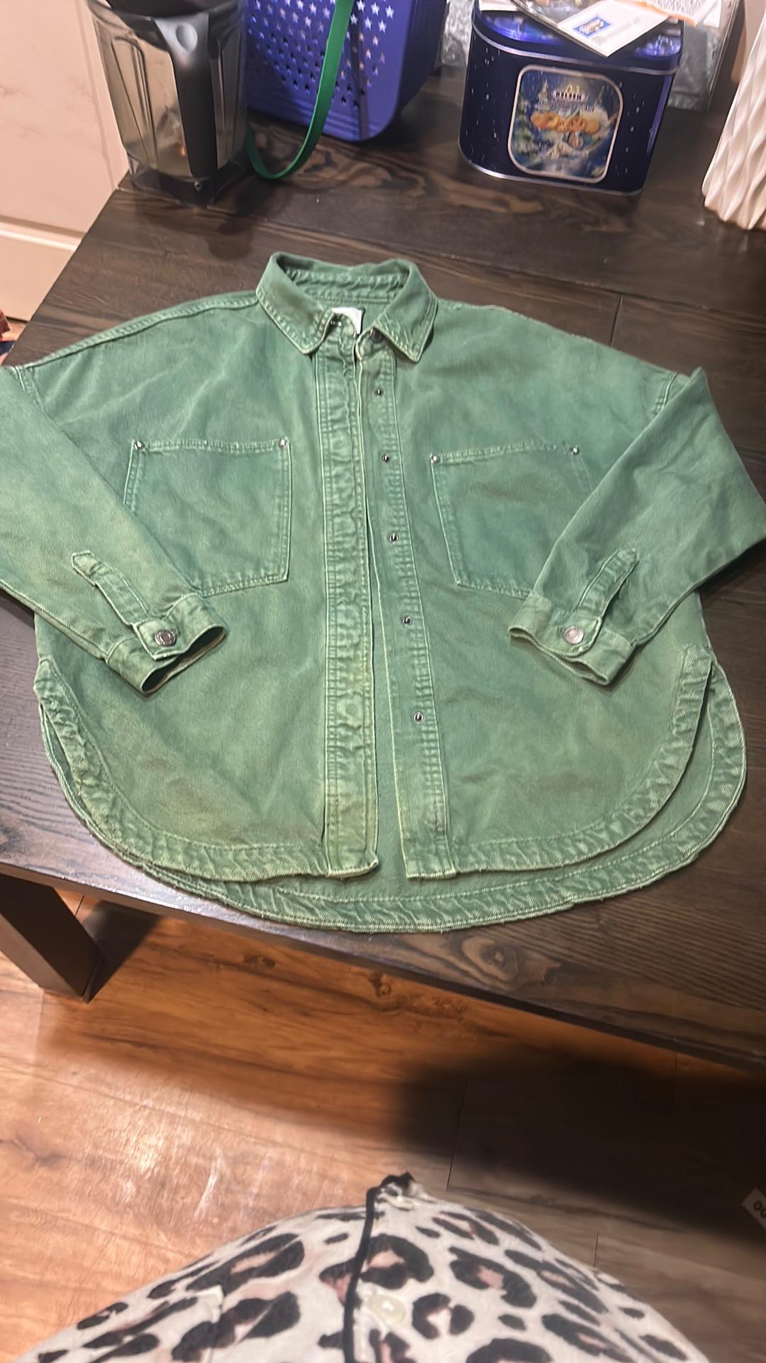 Casual style green shirt with front pockets on a dark wooden table, background with household objects and wooden floor.