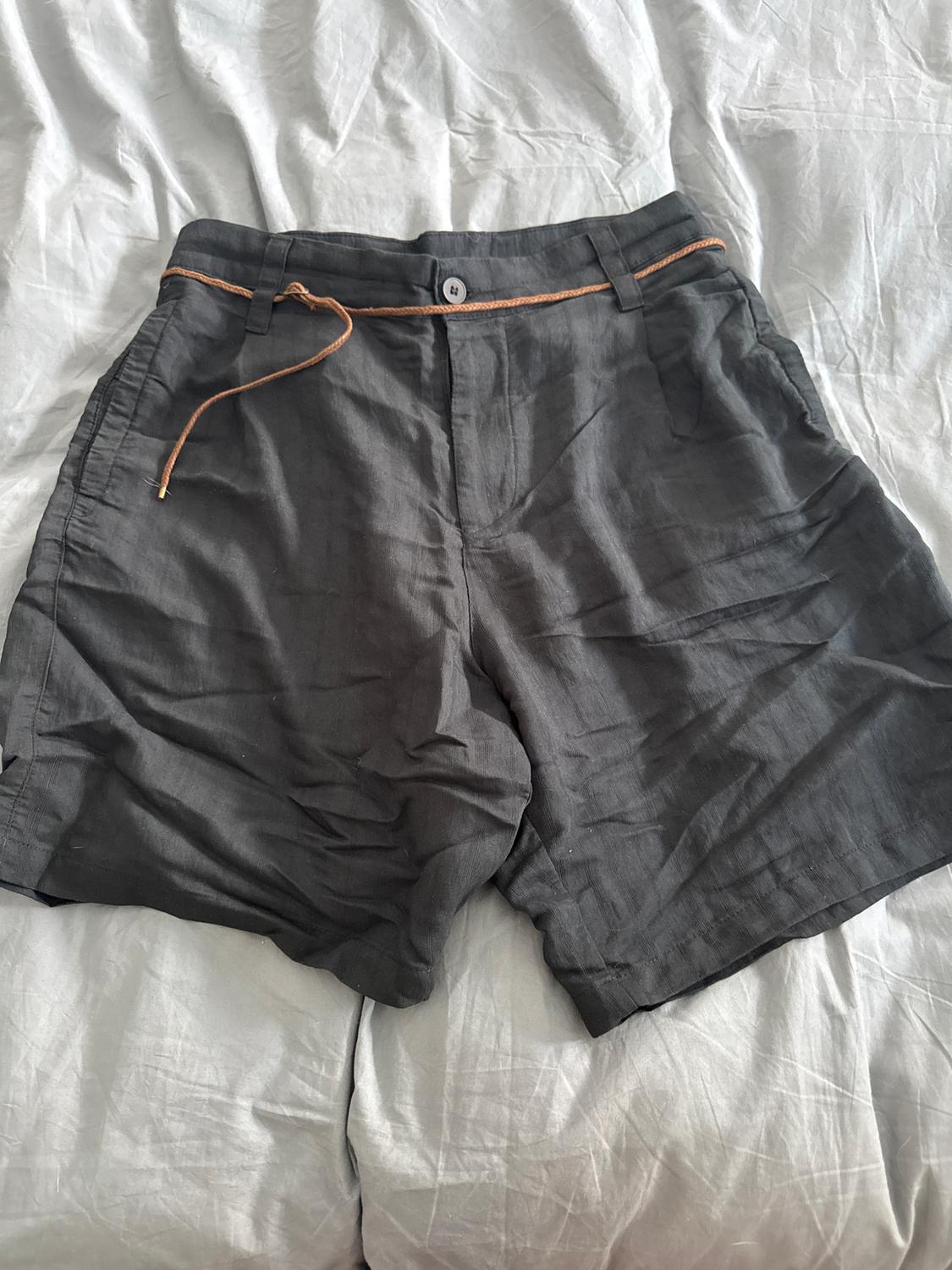 Grey crinkled linen shorts with a brown rope belt, on a bed with white sheets.