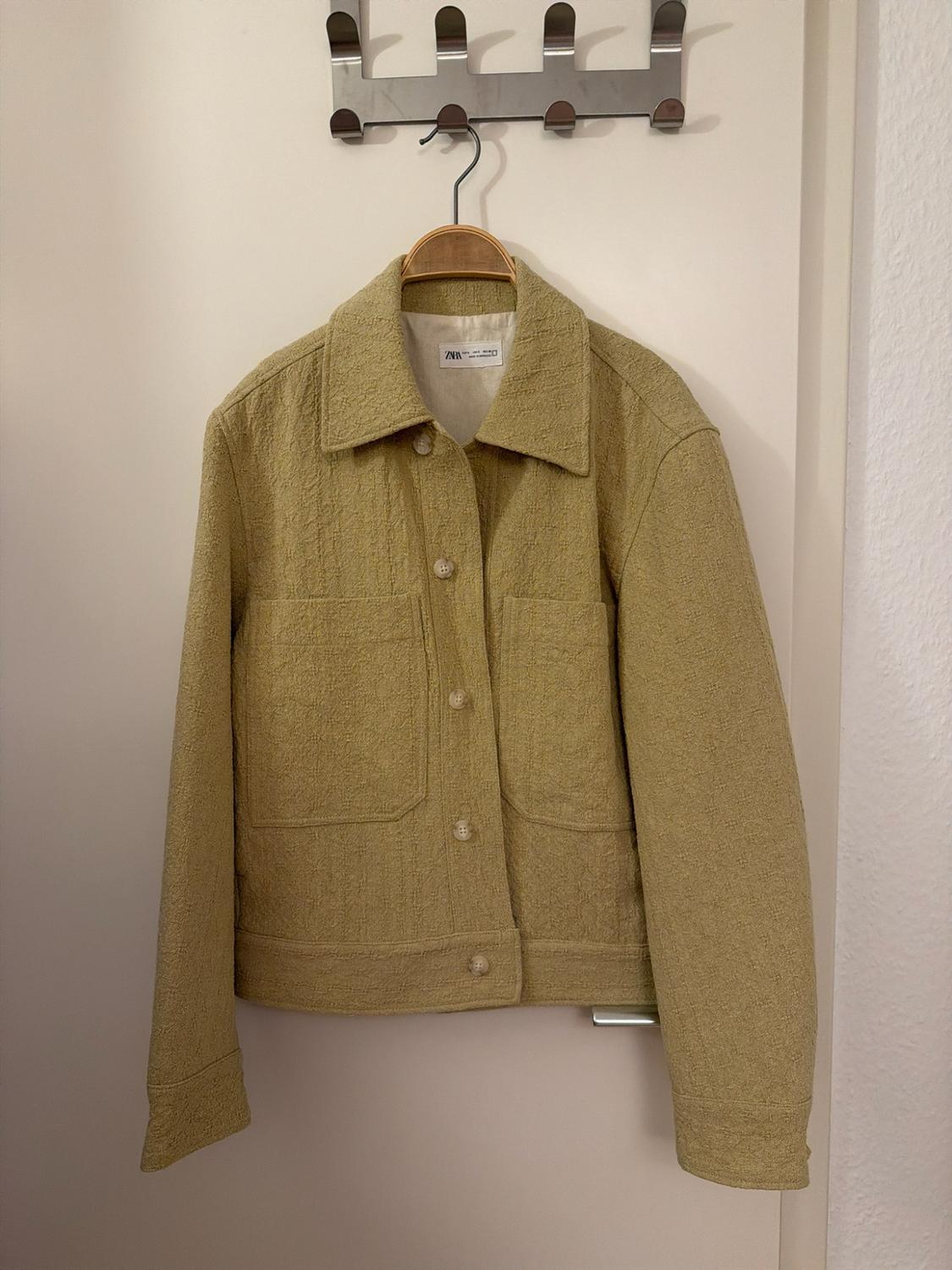 Textured light green jacket with buttons and pockets, hanging on a hanger on a white door. ZARA label visible.