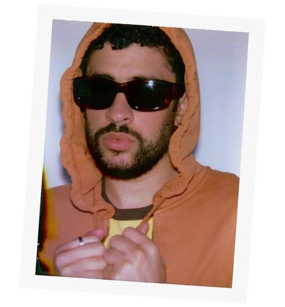 Hombre con lentes de sol y sudadera naranja con capucha, mirando a la cámara. Fondo blanco, estilo relajado y moderno.