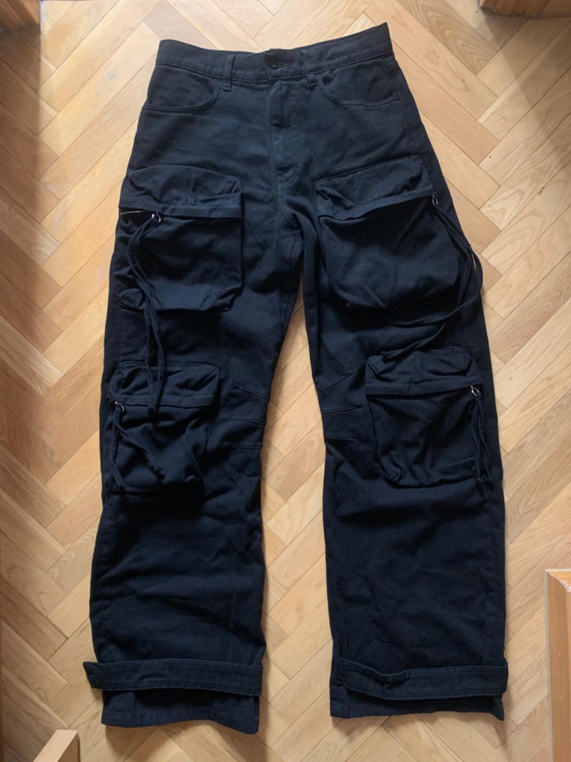Black cargo trousers with large pockets and straps, photographed on a herringbone wooden floor. Urban and relaxed style.