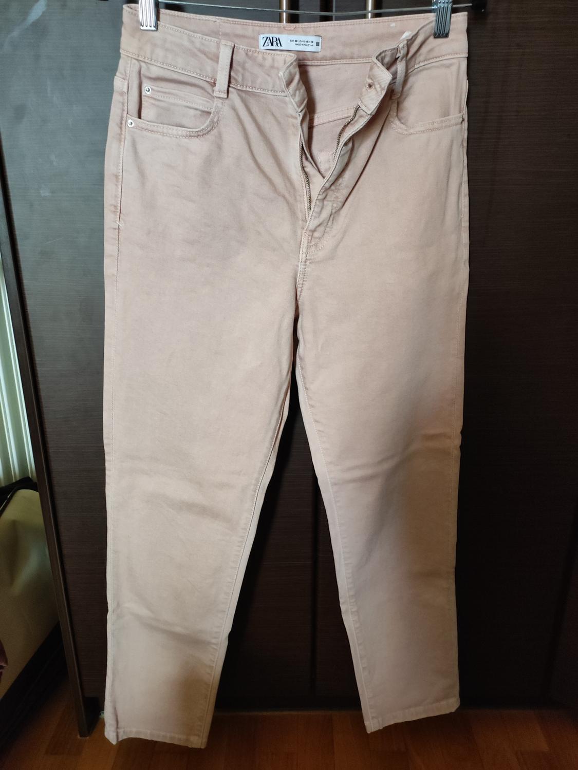Beige trousers hanging on a hanger, with an open zip and visible Zara tag. Dark wood background.