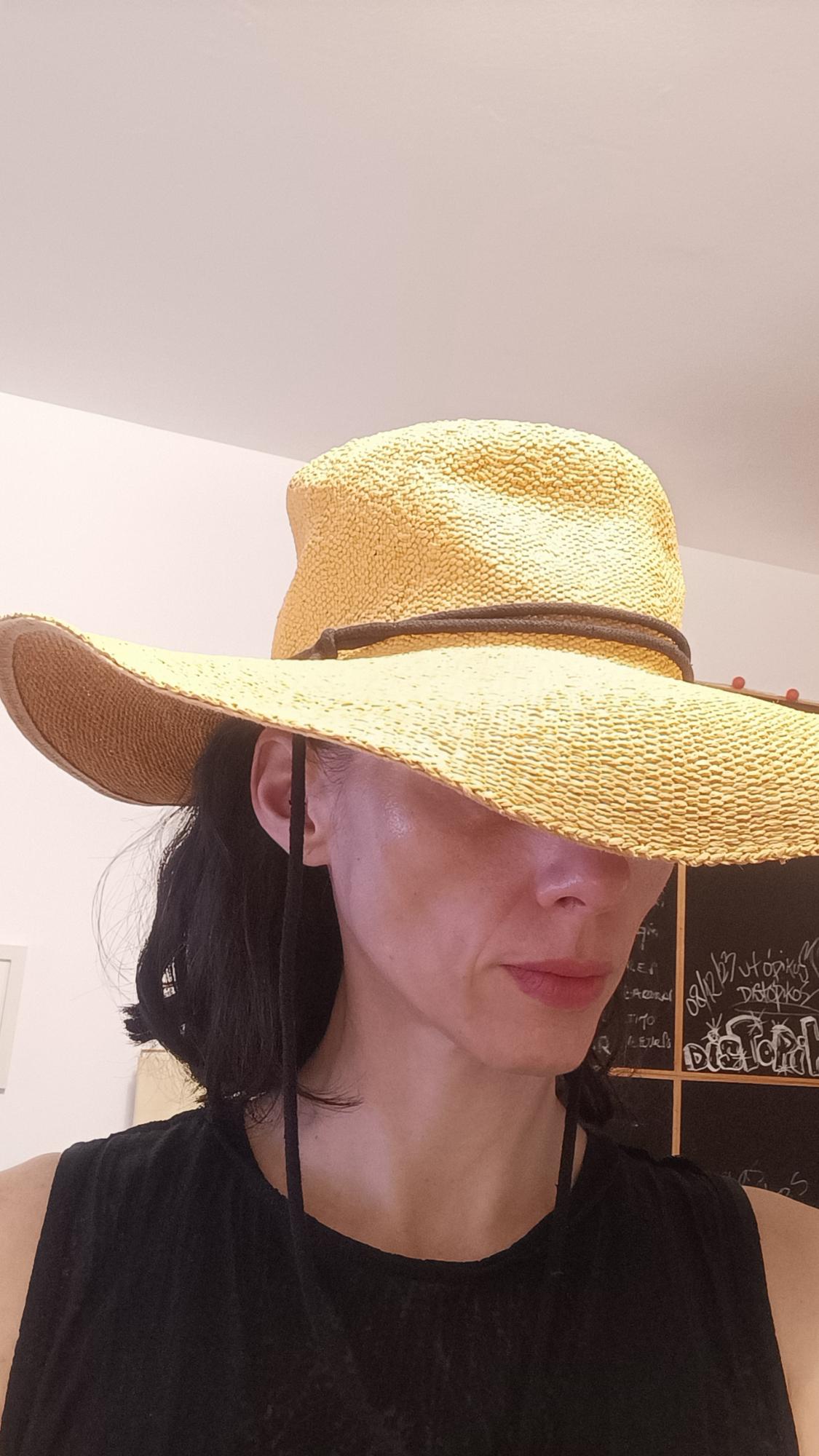 Woman in a black top and large yellow hat obscuring her eyes. Chalkboard in the background with text "08/12/23 utópikos distópikos".