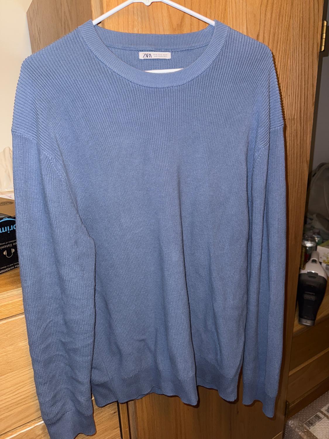 Blue knit sweater hanging on a white hanger in front of a wooden wardrobe. Visible tag with the text "ZARA".