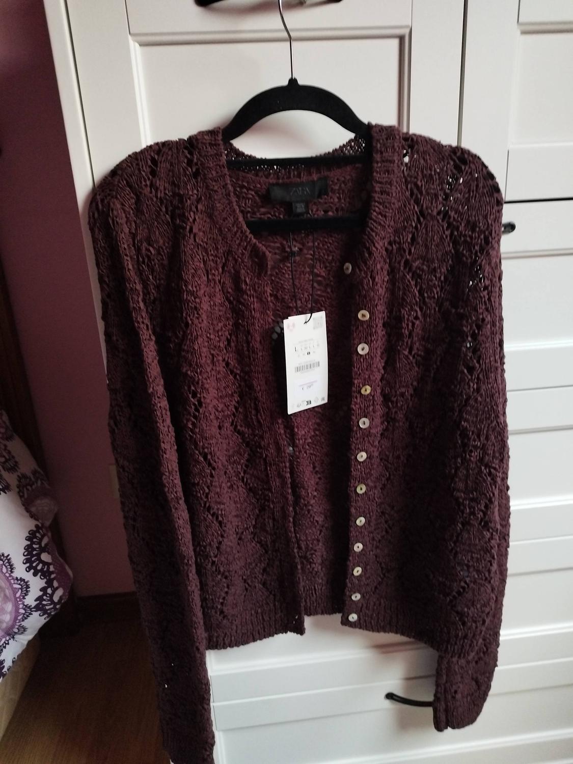 Brown knitted cardigan hanging on a black hanger, with visible tag, against a white furniture background.
