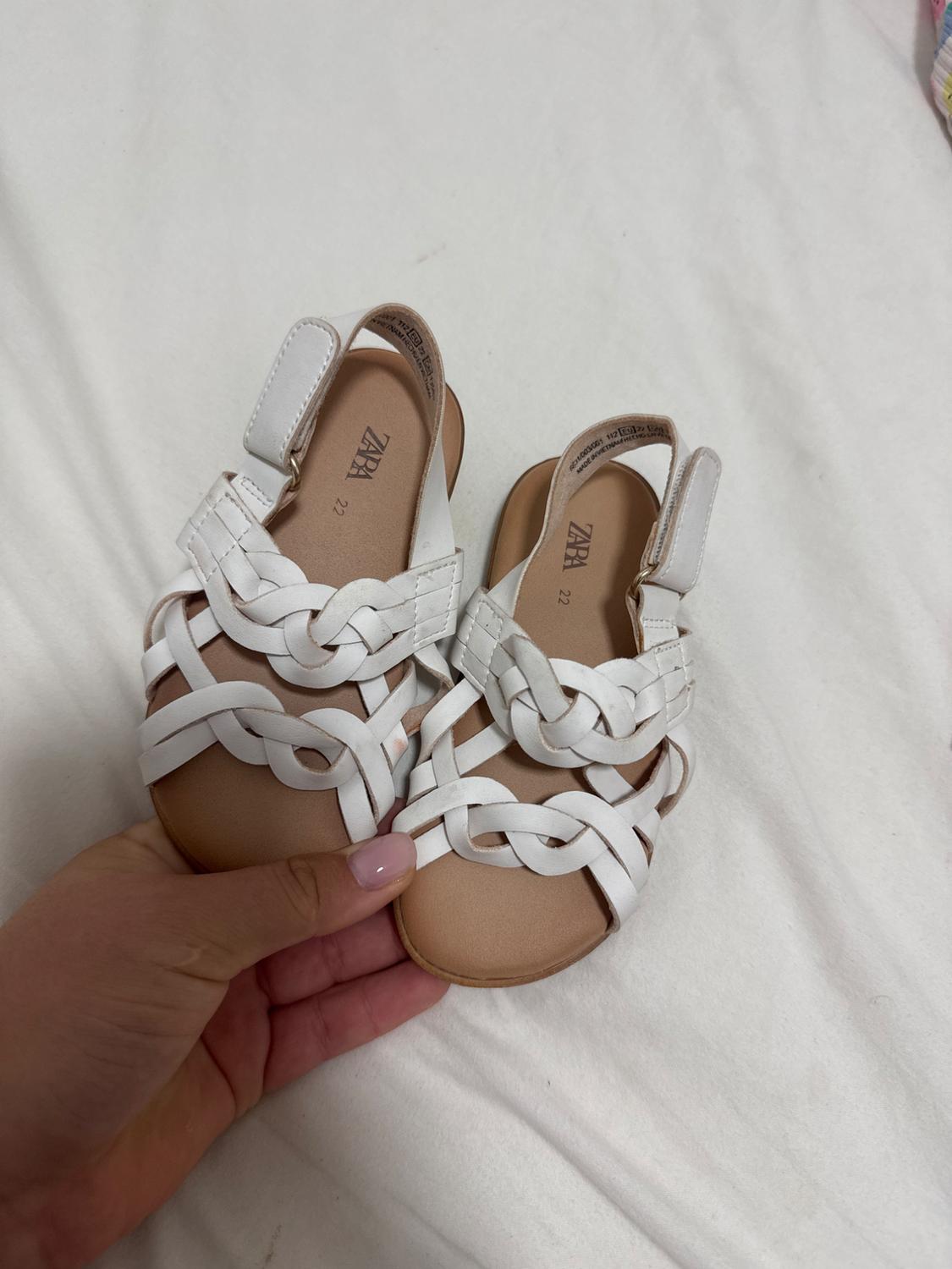 Hand holding white sandals with intertwined straps against a white fabric background. Visible text: ZARA 22.