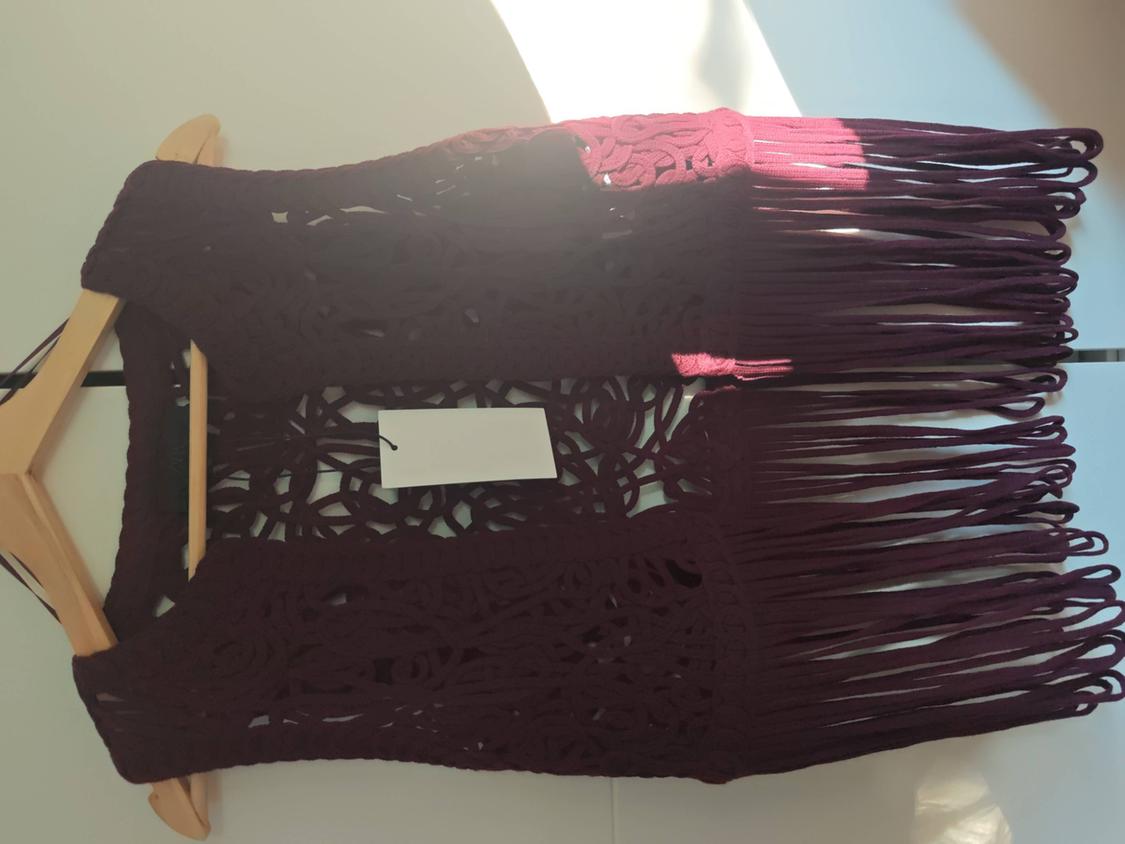 Burgundy open-knit top with long fringing at the hem, hanging on a wooden hanger against a white background with natural light and shadows.