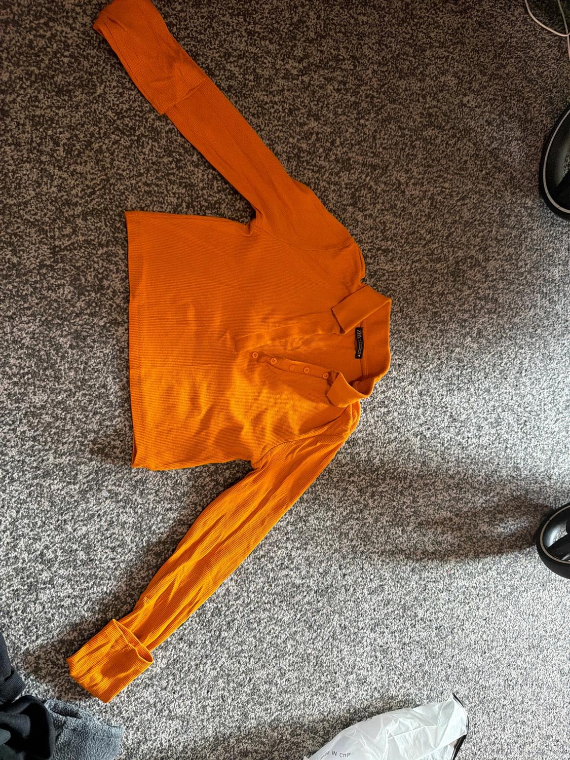 Orange ribbed long-sleeved cropped top with a polo neck on a grey carpet. A white bag bearing the text IN CHINA can be seen.