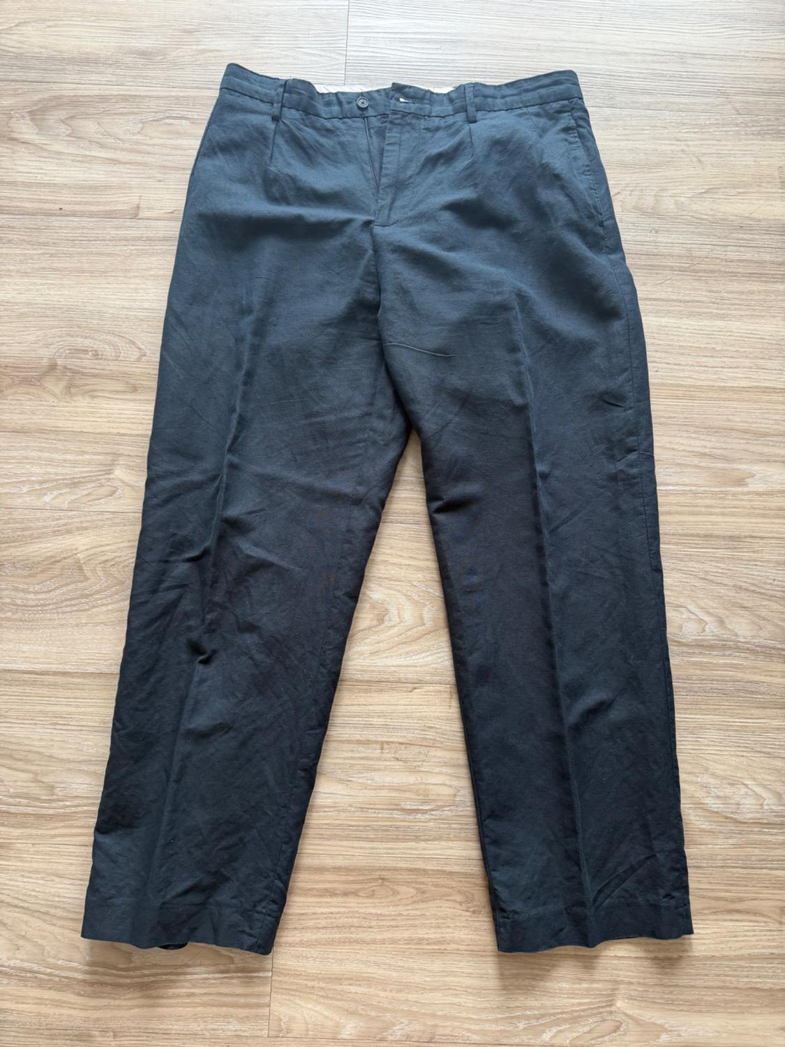 Dark, straight-cut trousers with button fastening, casual and relaxed style, photographed flat on a light wooden floor.