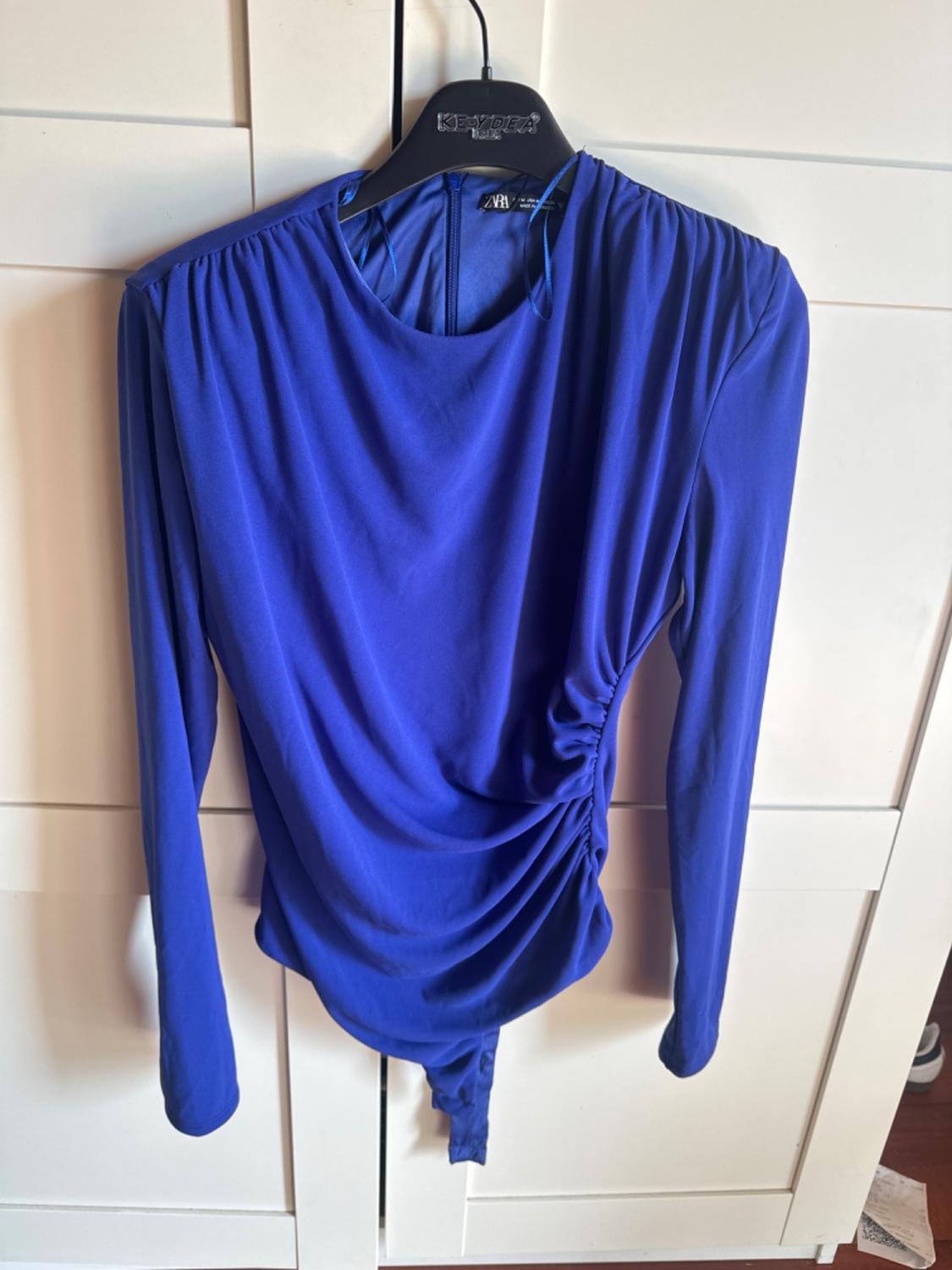 Electric blue long-sleeved bodysuit with side ruching, hanging on a black hanger in front of a white wardrobe.