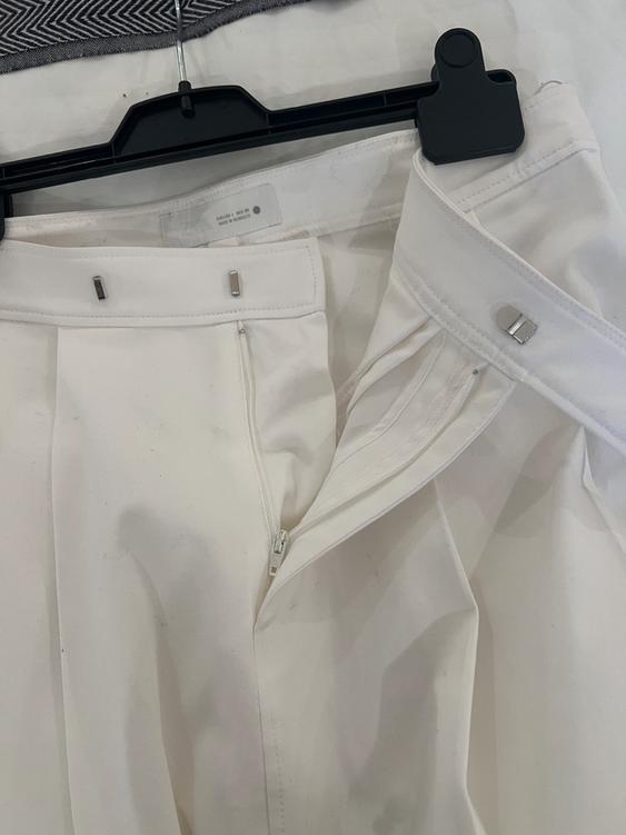 White trousers hanging on a black hanger, with a zip fly and button fastening. Fabric background with a herringbone pattern.