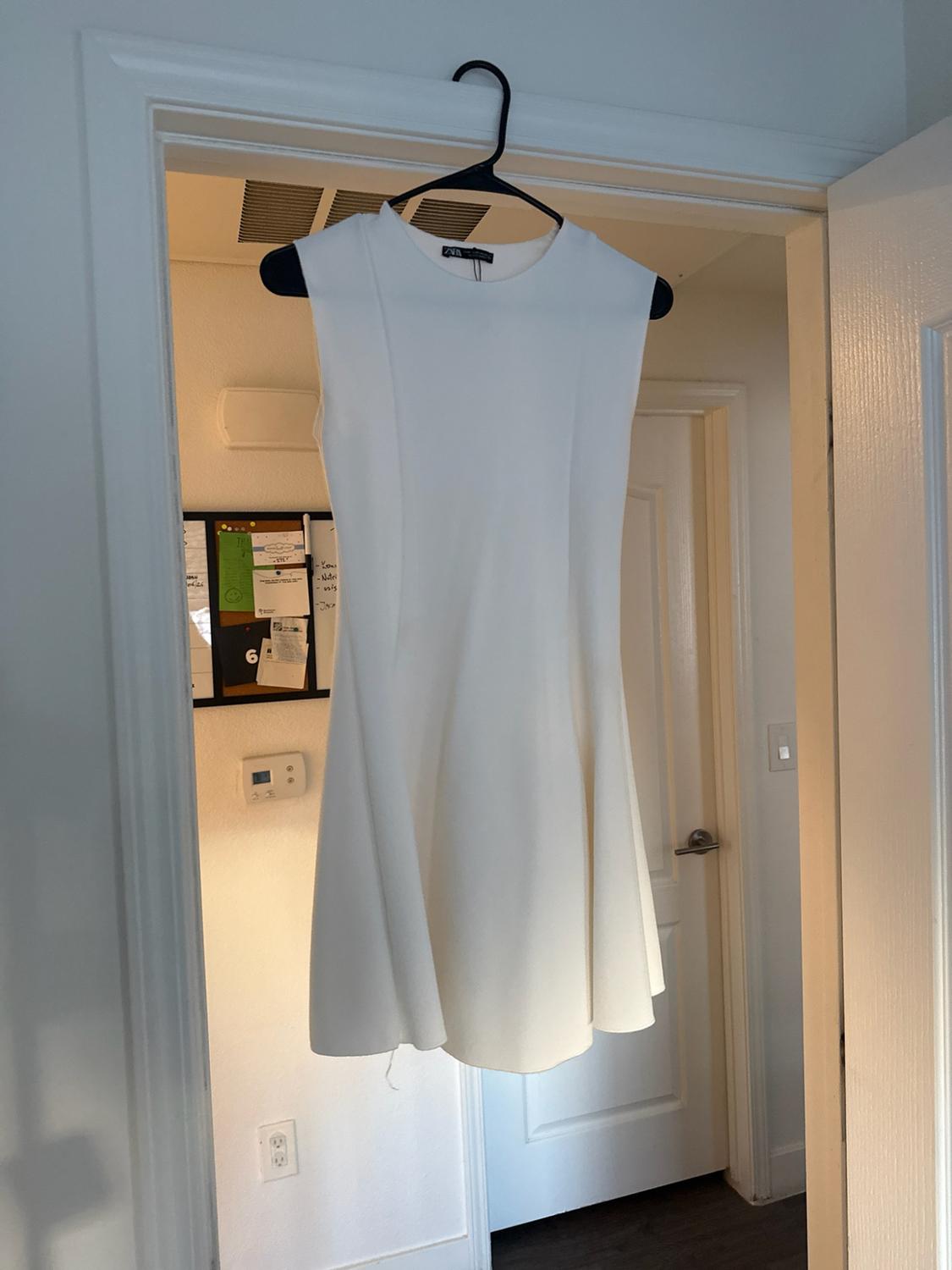 Short white sleeveless dress with a flared skirt, hanging on a black hanger in a doorway indoors.
