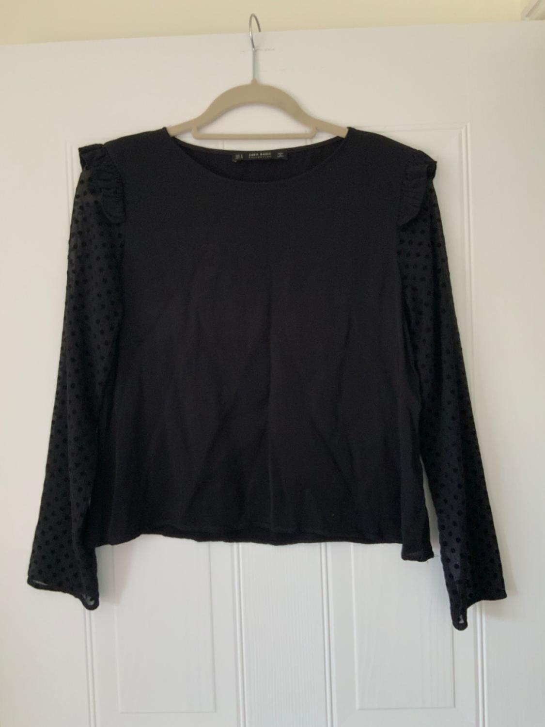 Front view of a black blouse with long transparent polka dot sleeves and ruffles on the shoulders.