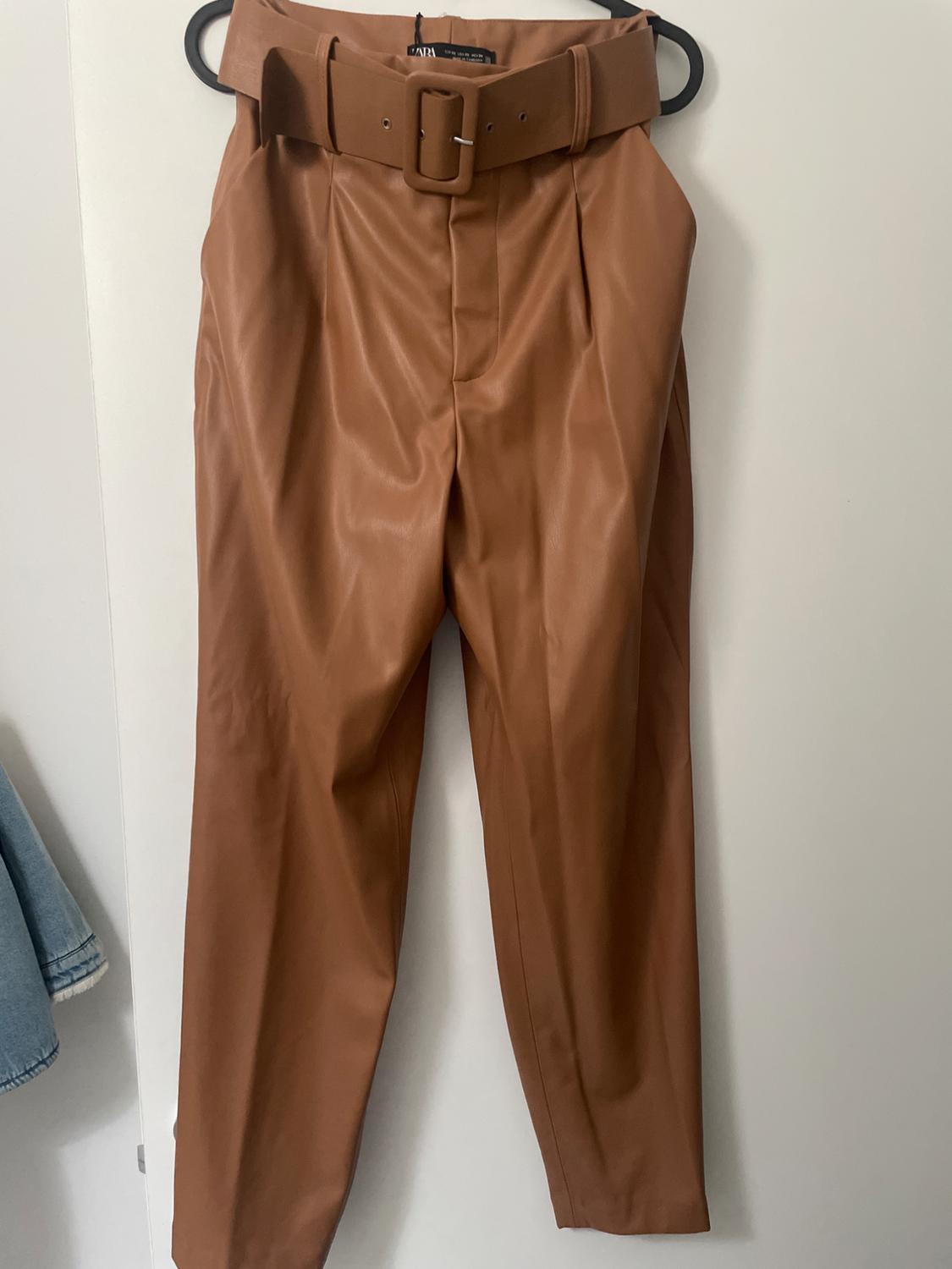 Brown trousers hanging on a hanger, with a matching wide belt and pleats at the top. White background.