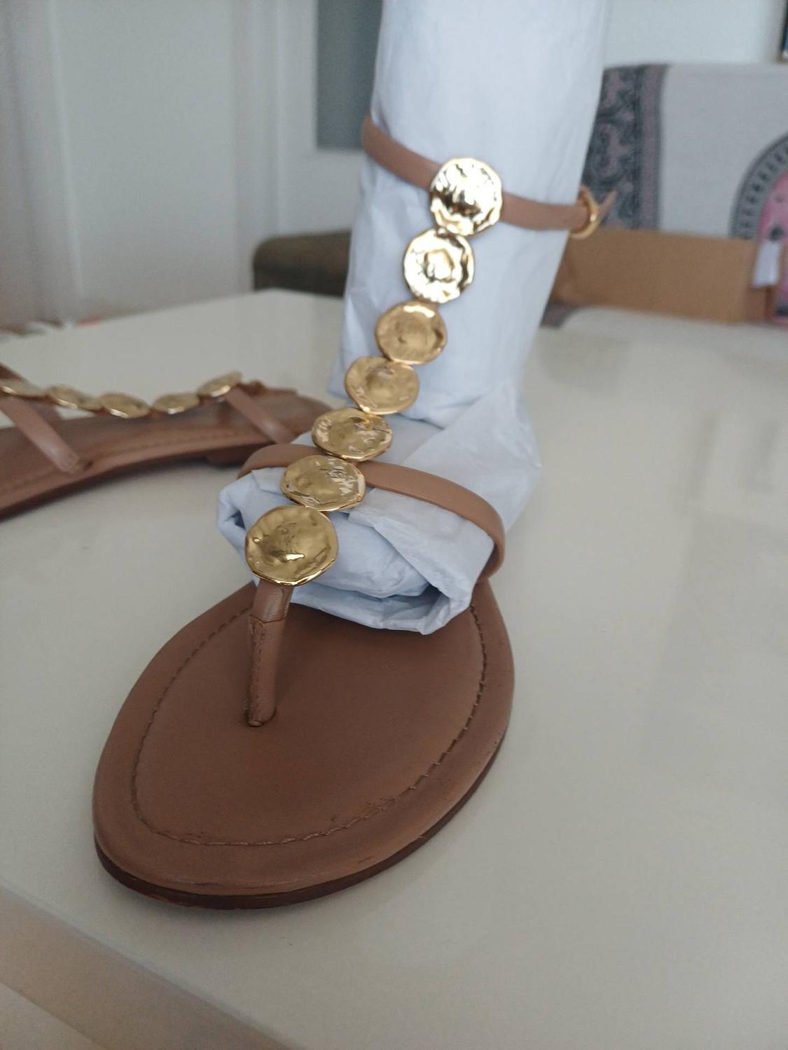Brown flat sandals with straps and gold discs. One of them is stuffed with white paper on a light, shiny surface.