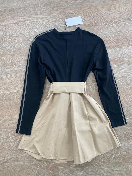 Dress on hanger on wooden floor. Long-sleeved black top, pleated beige skirt. White tag hanging.