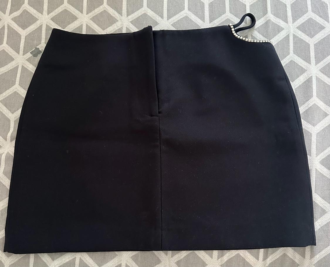 Black mini skirt against a geometric background. It features a central seam and a slit detail with a strap and rhinestones at the top right.