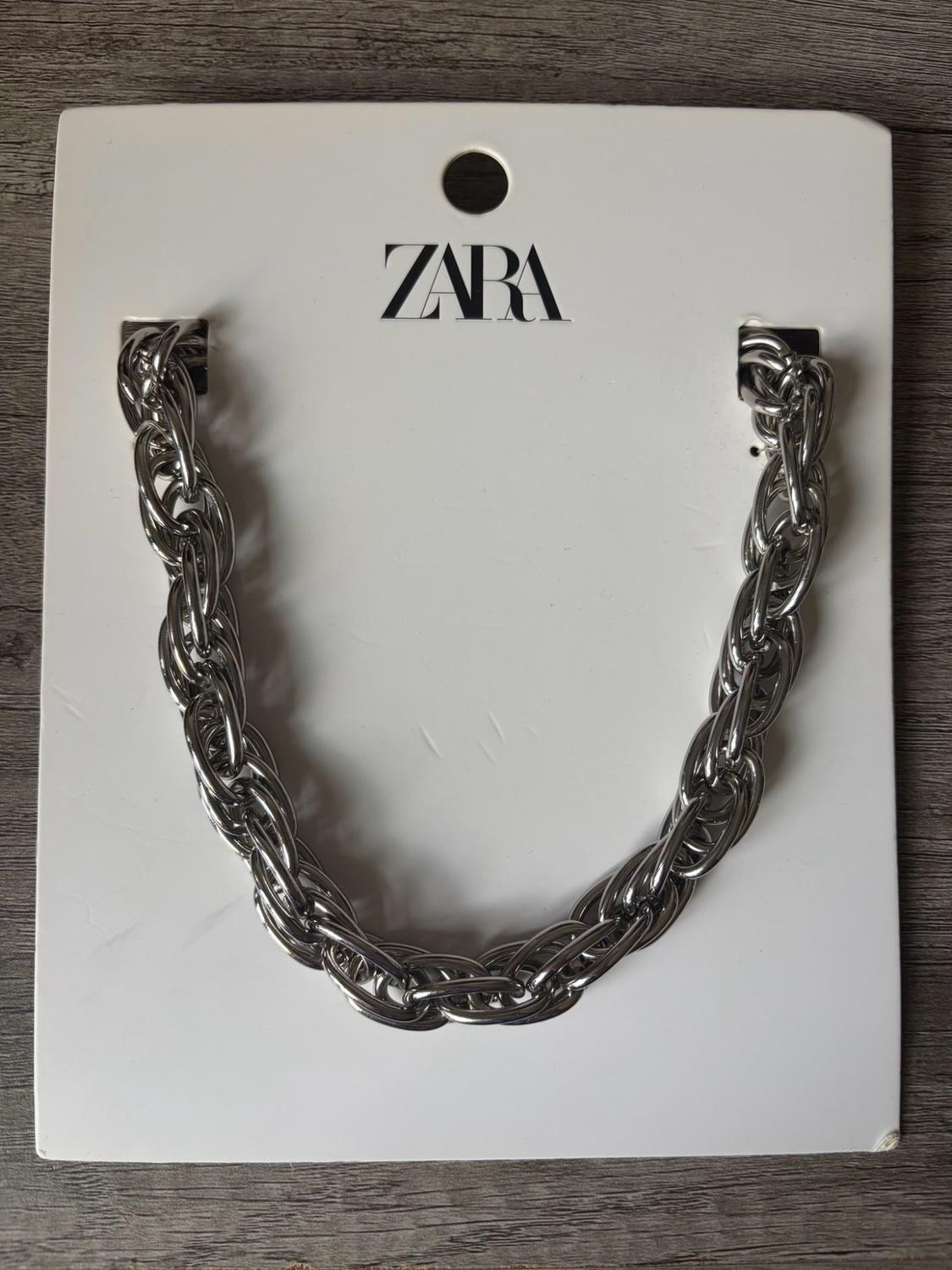 Chunky silver chain necklace on a white cardboard display with the text ZARA printed on it, on a wooden surface.