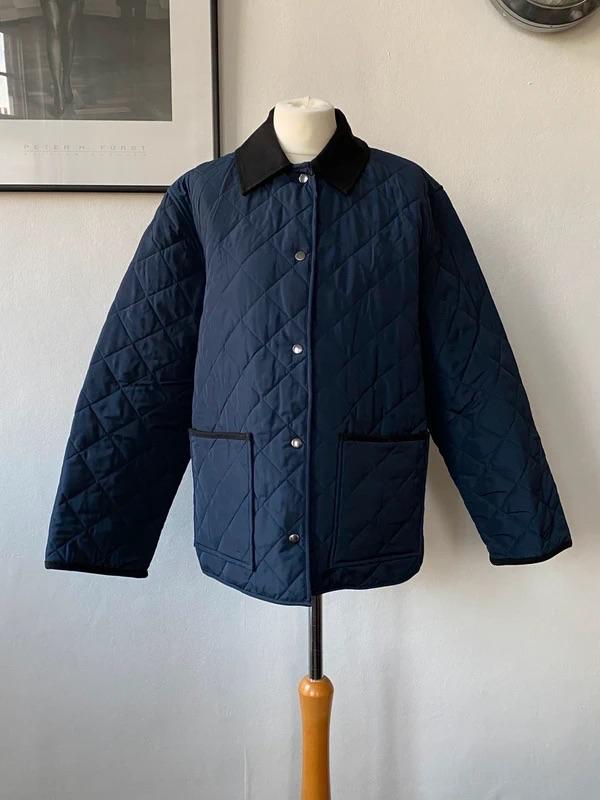 Blue quilted jacket with a black collar on a mannequin, white background. Picture on the wall and partially visible clock.