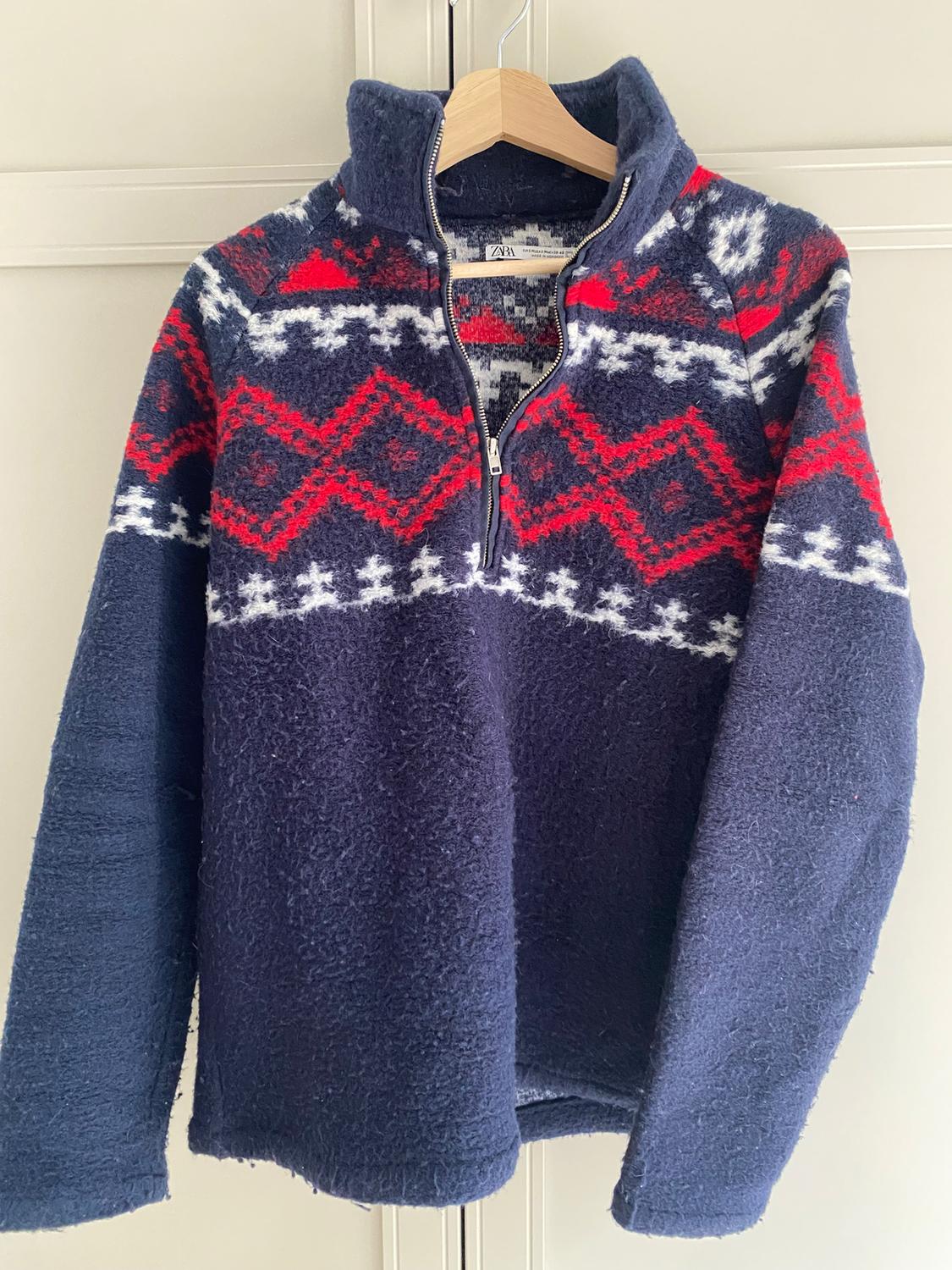 Blue wool jumper with zip, red and white geometric design on the chest and shoulders, hanging on a wooden hanger.